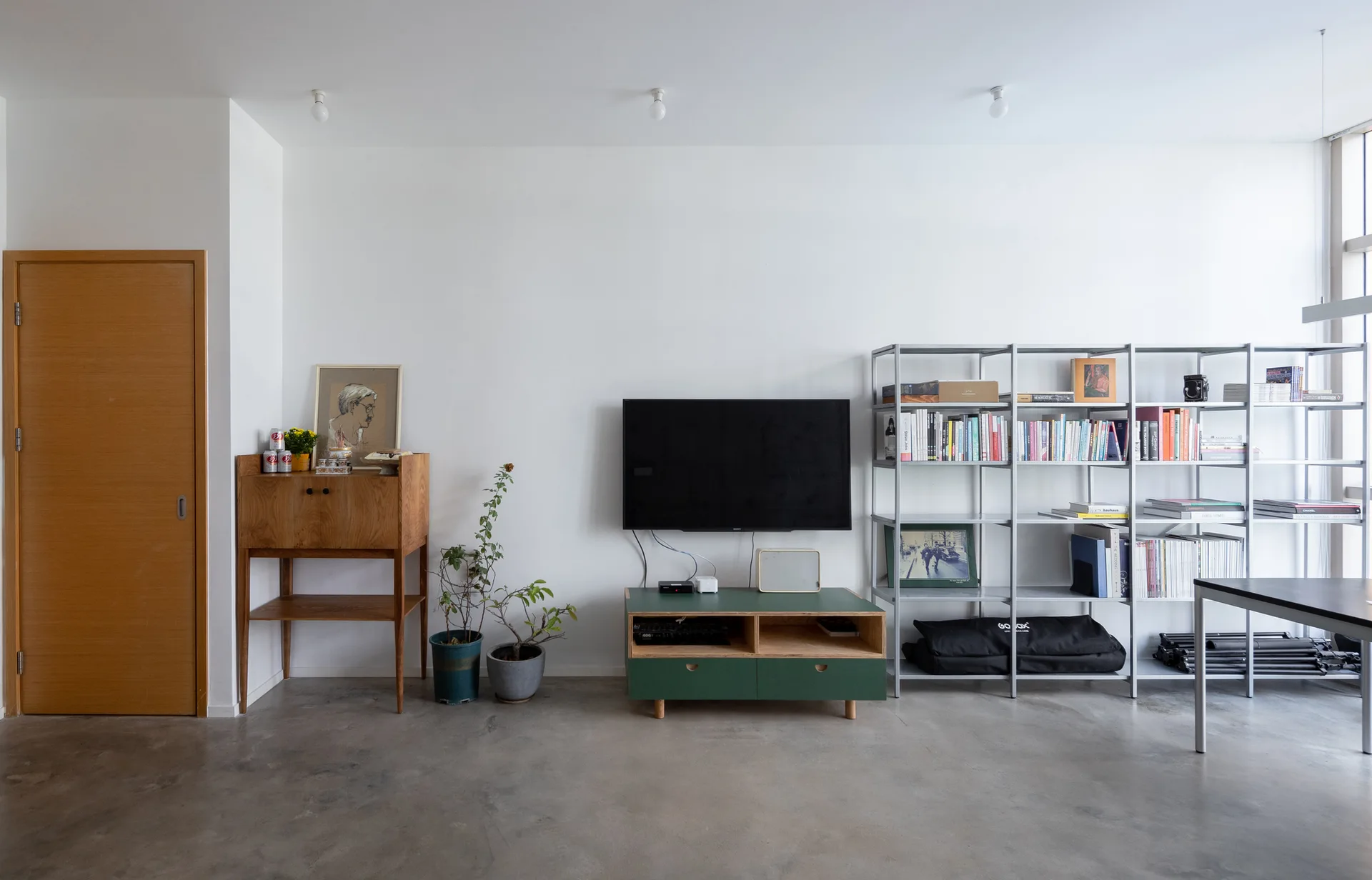 A minimalist living room features a green media console below a wall-mounted television, flanked by a wooden cabinet and tall grey metal shelving.