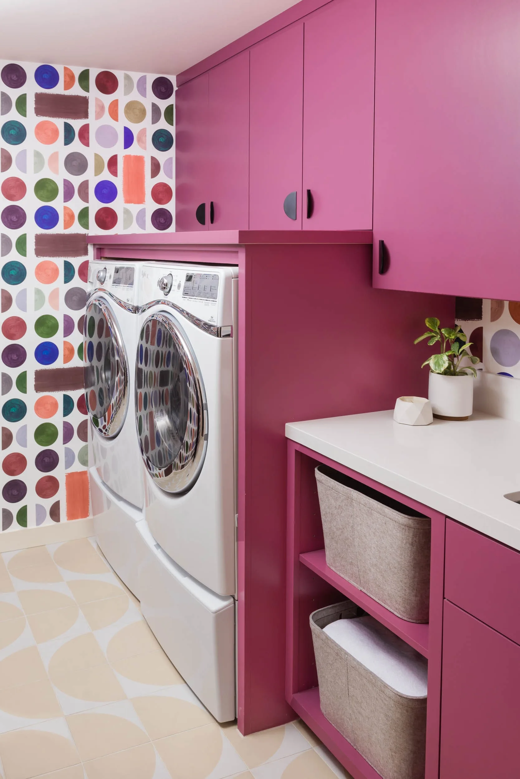 A laundry room features custom magenta cabinets surrounding a white washer and dryer next to a white countertop and colorful geometric wallpaper.