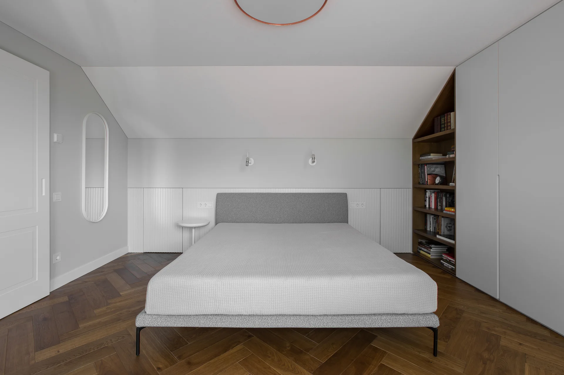 A minimalist attic bedroom features a grey upholstered bed, dark wood herringbone flooring, and built-in wardrobes alongside a custom triangular bookshelf.