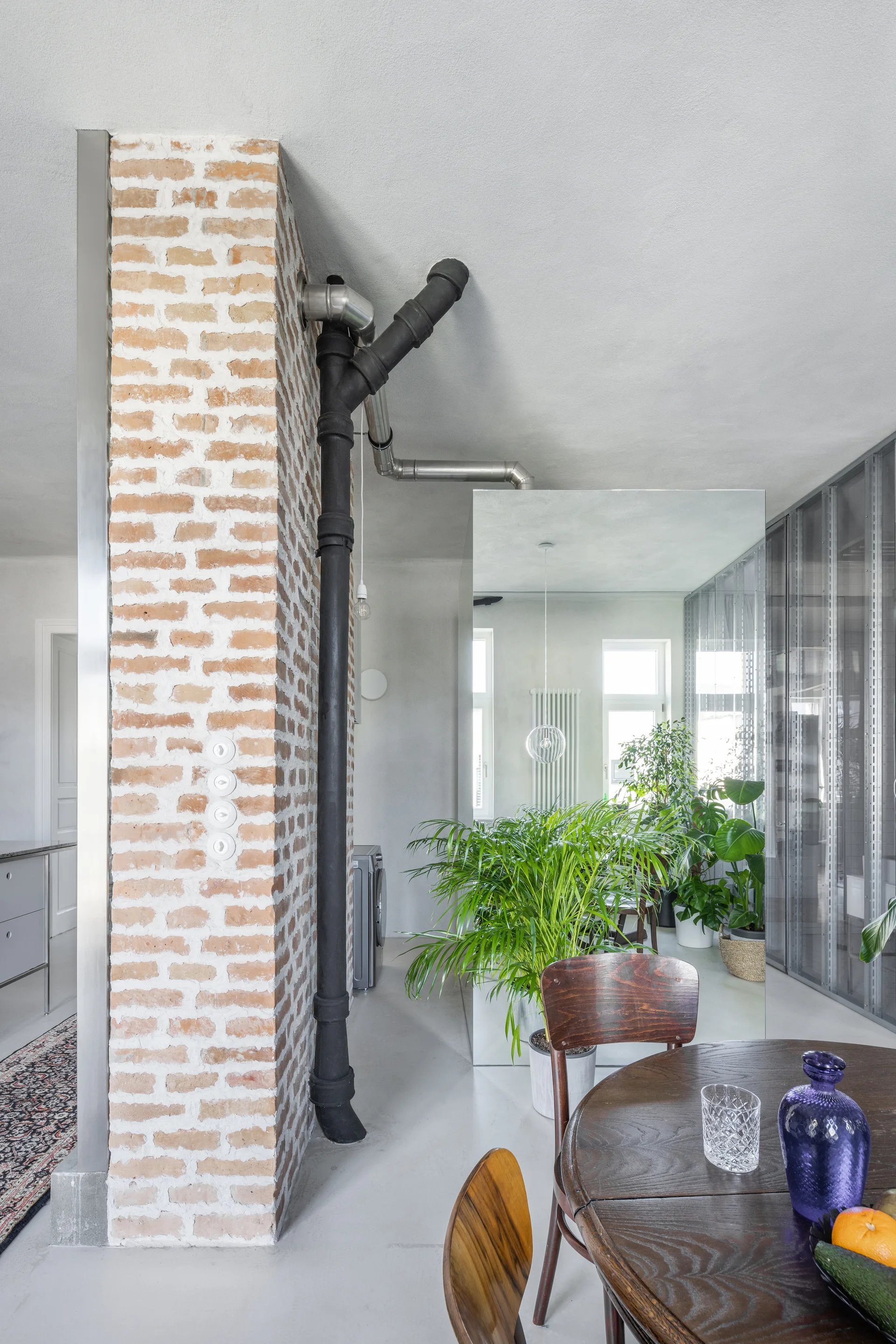 An industrial dining area features a dark wood table, an exposed brick column with black plumbing pipes, and a large mirror reflecting potted plants.