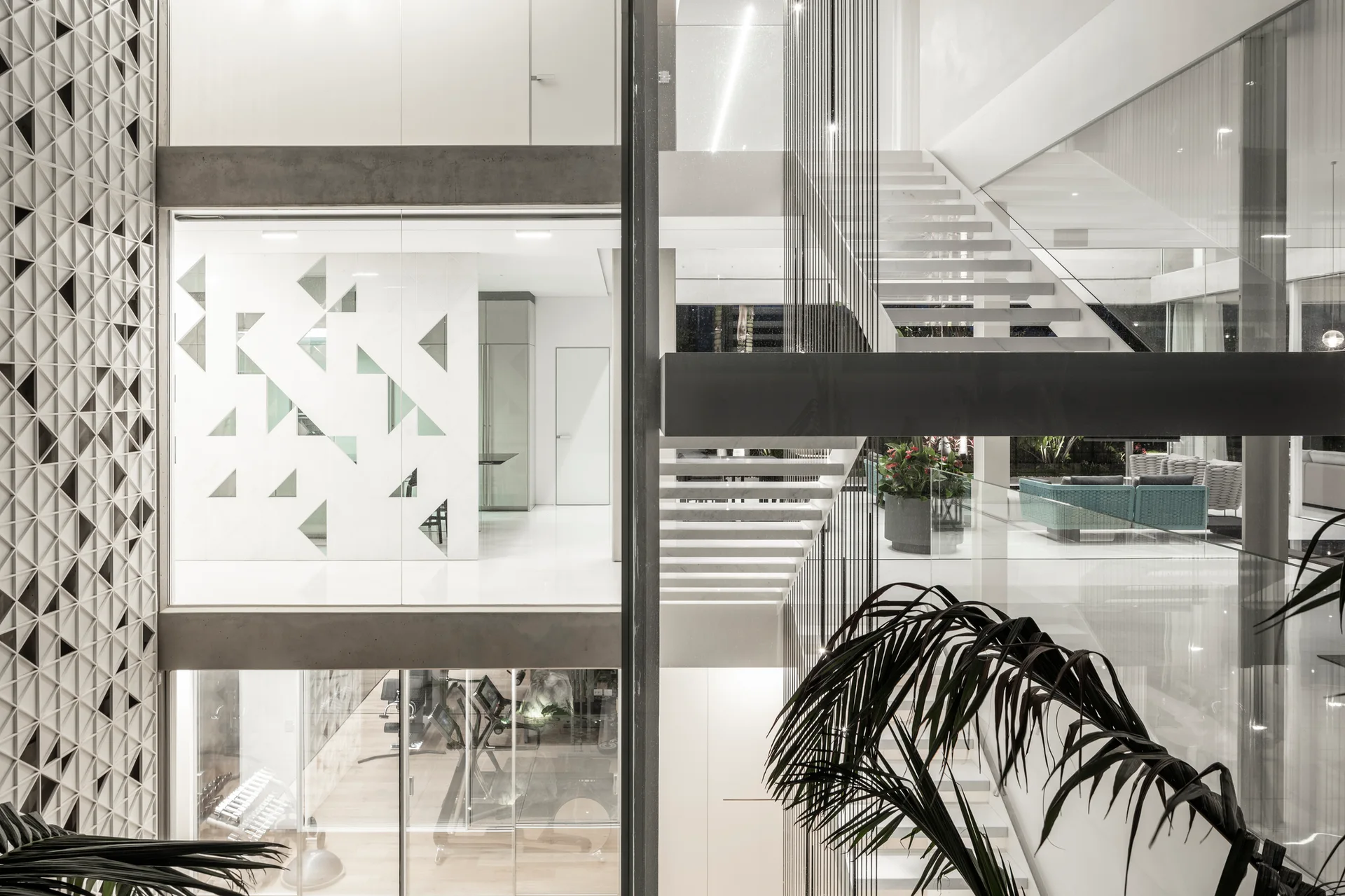A multi-level home interior featuring a central floating staircase with glass railings, concrete beams, and a white geometric lattice wall screen.