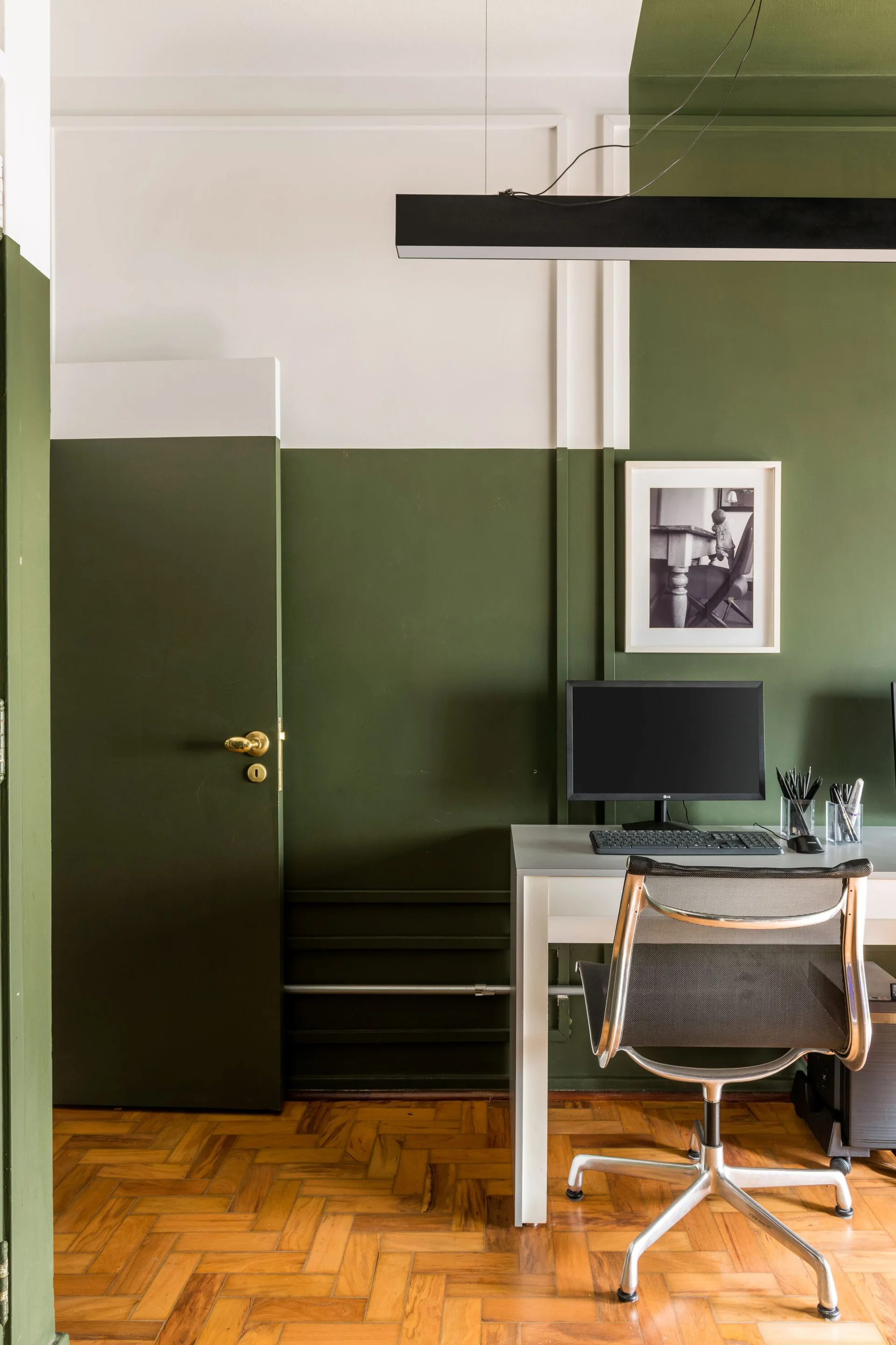 A minimalist home office with a white desk, mesh chair, and parquet wood floor against a wall color-blocked in olive green and white.