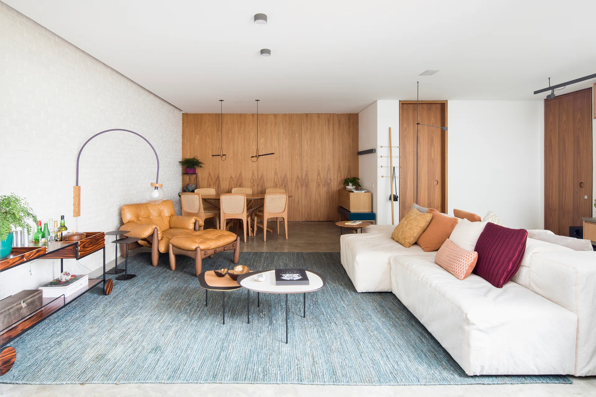 An open-plan living room features a cream sectional sofa on a blue textured rug, a tan leather armchair, and a warm wood-paneled accent wall.