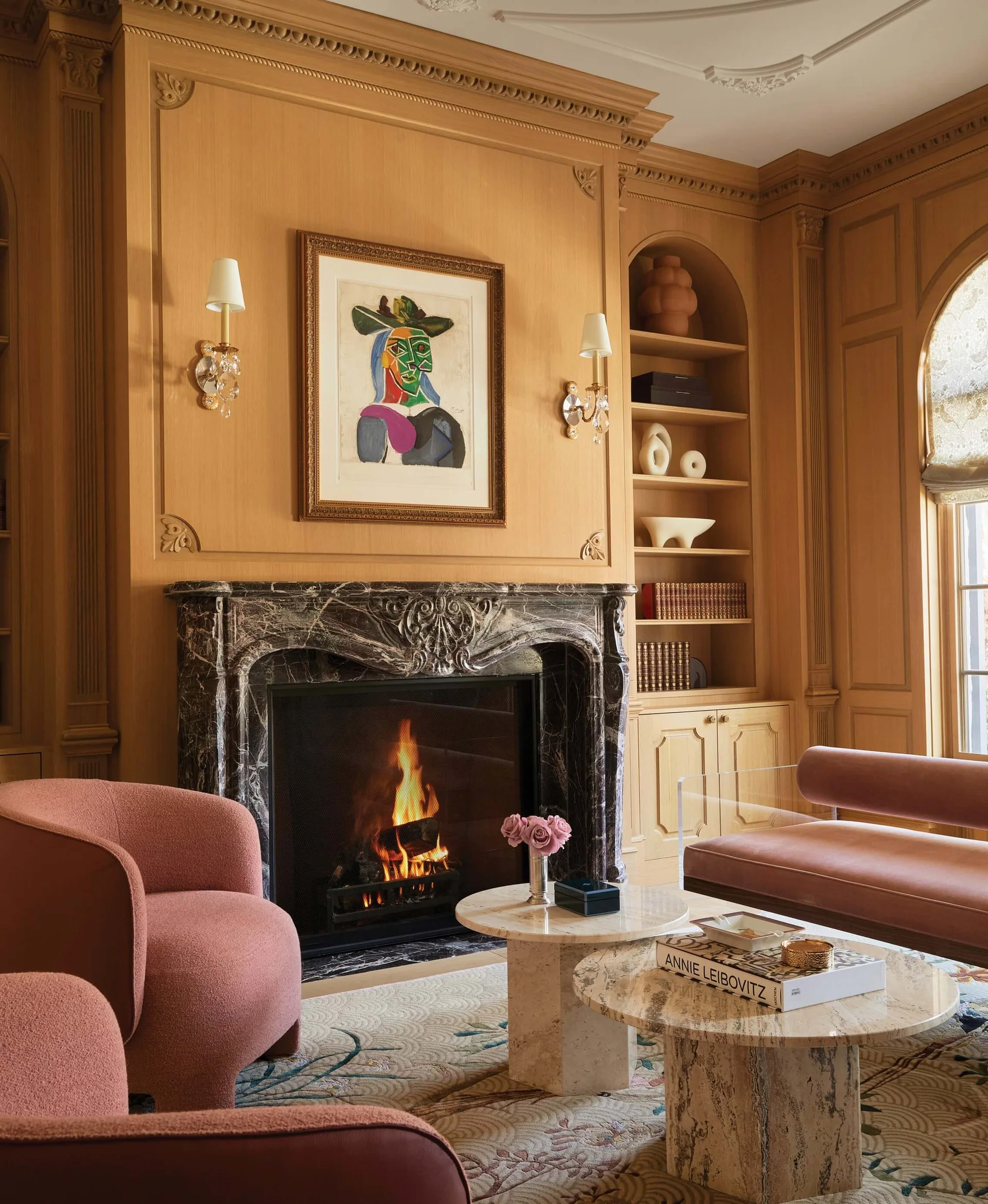 A living room features warm wood-paneled walls, an ornate dark marble fireplace with a burning fire, pink armchairs, and tiered stone coffee tables.