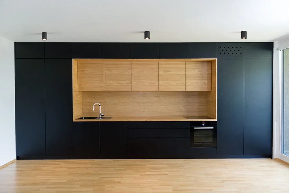 A wall of matte black minimalist kitchen cabinets featuring a recessed central light wood niche with a sink and countertop over light wood flooring.