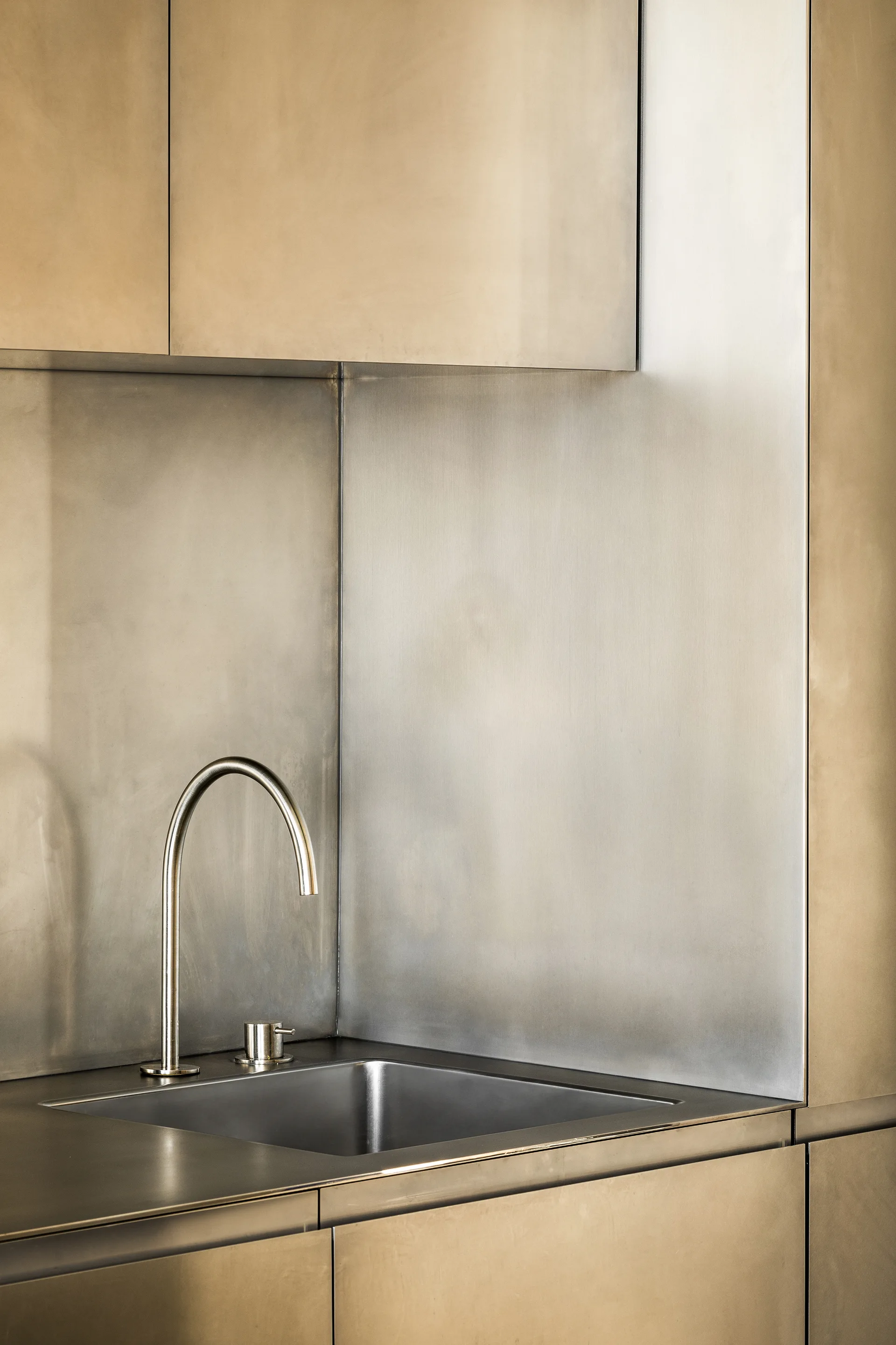 A minimalist kitchen sink with a curved gooseneck faucet is surrounded by brushed bronze cabinetry and a seamless metal backsplash.
