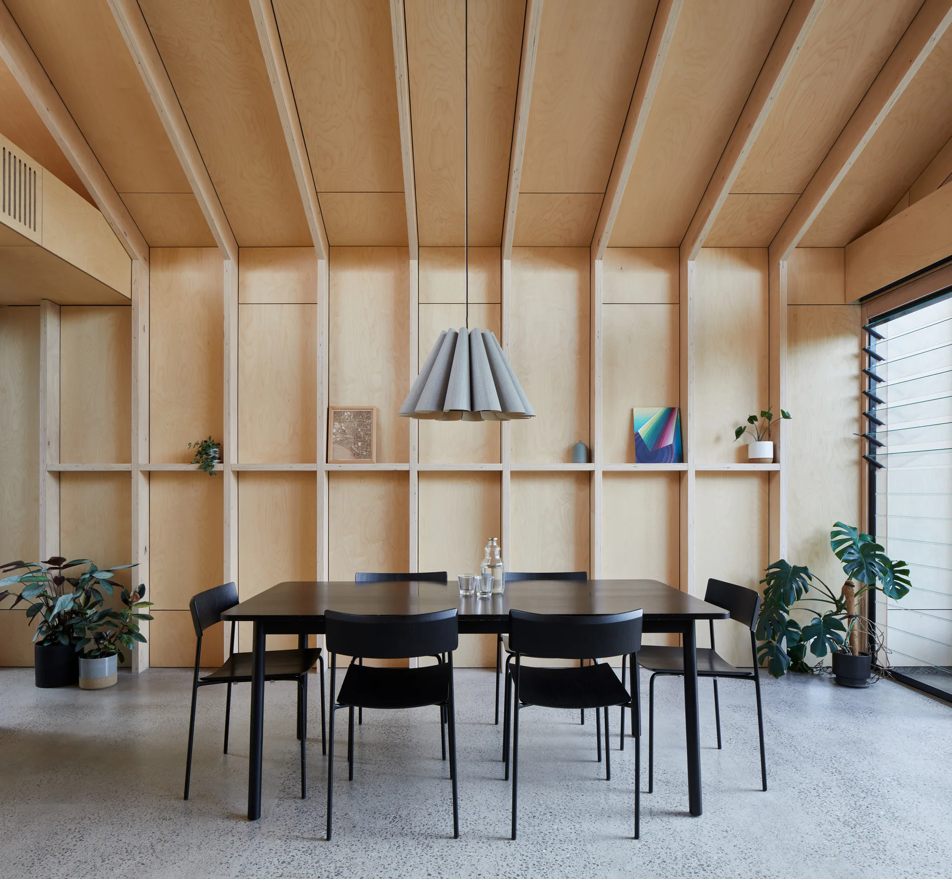 Minimalist dining room with a black table and chairs against a light plywood paneled wall beneath a pleated pendant light.