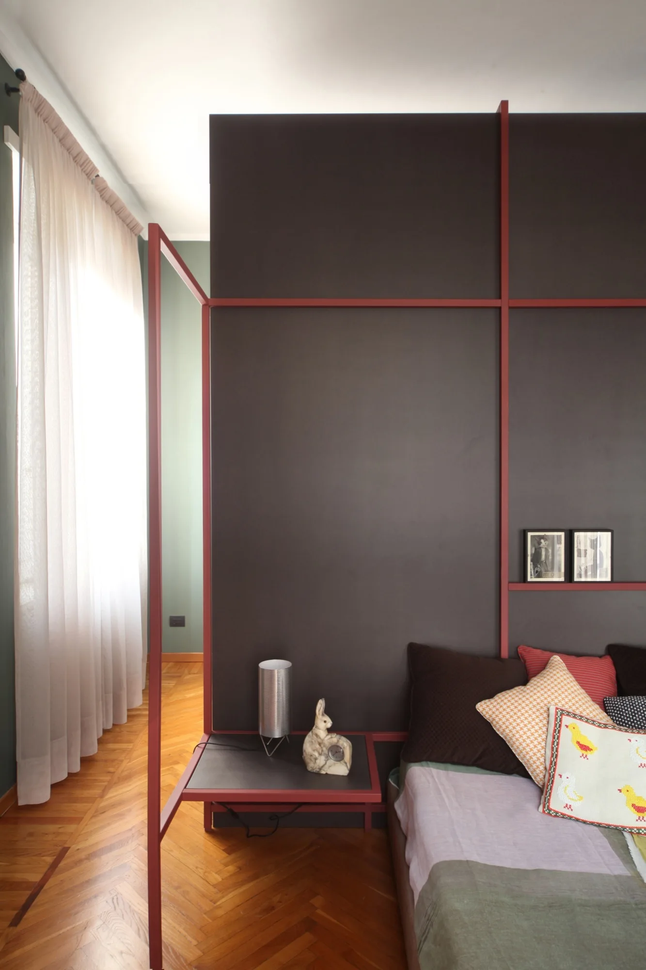 A modern bedroom features a dark gray partition wall with a red metal frame, sheer curtains, and a herringbone parquet wood floor.