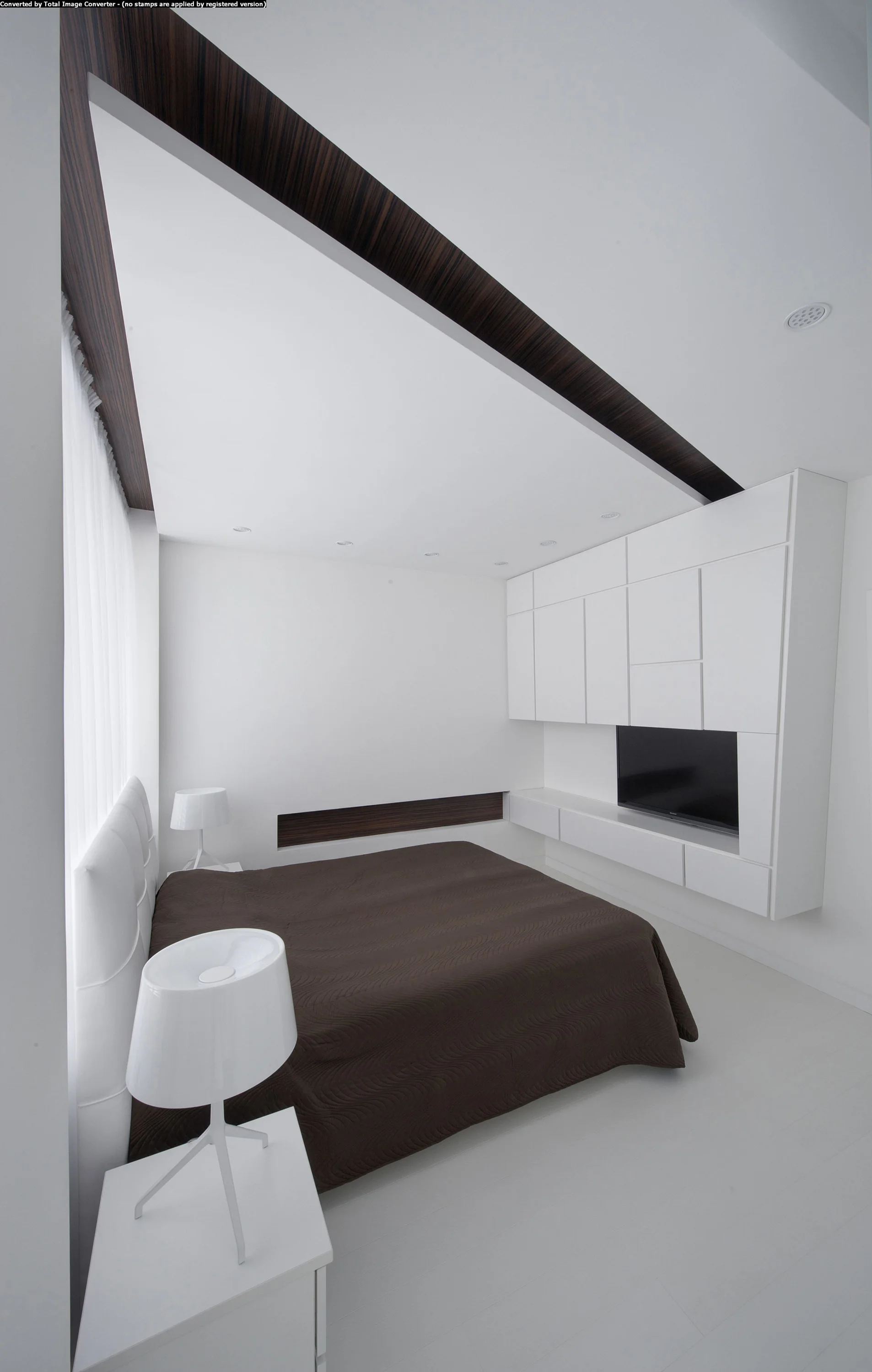 A minimalist white bedroom features a dark brown bed, white modern lamps, and custom built-in cabinetry beneath a diagonal dark wood ceiling accent.