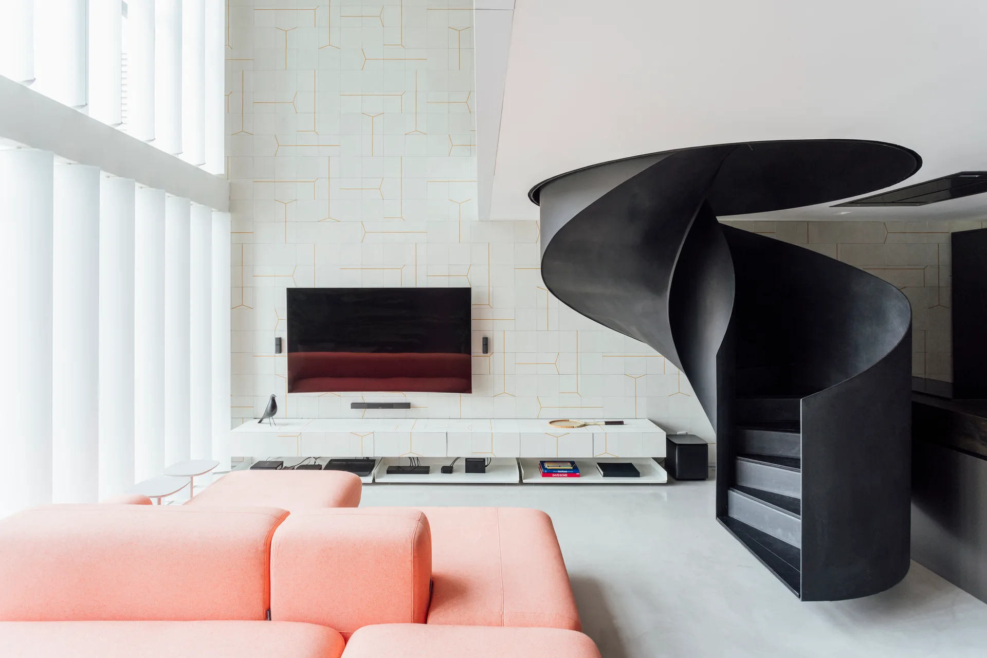 A minimalist living room features a salmon pink modular sofa, a prominent black spiral staircase, and a white tiled media wall.