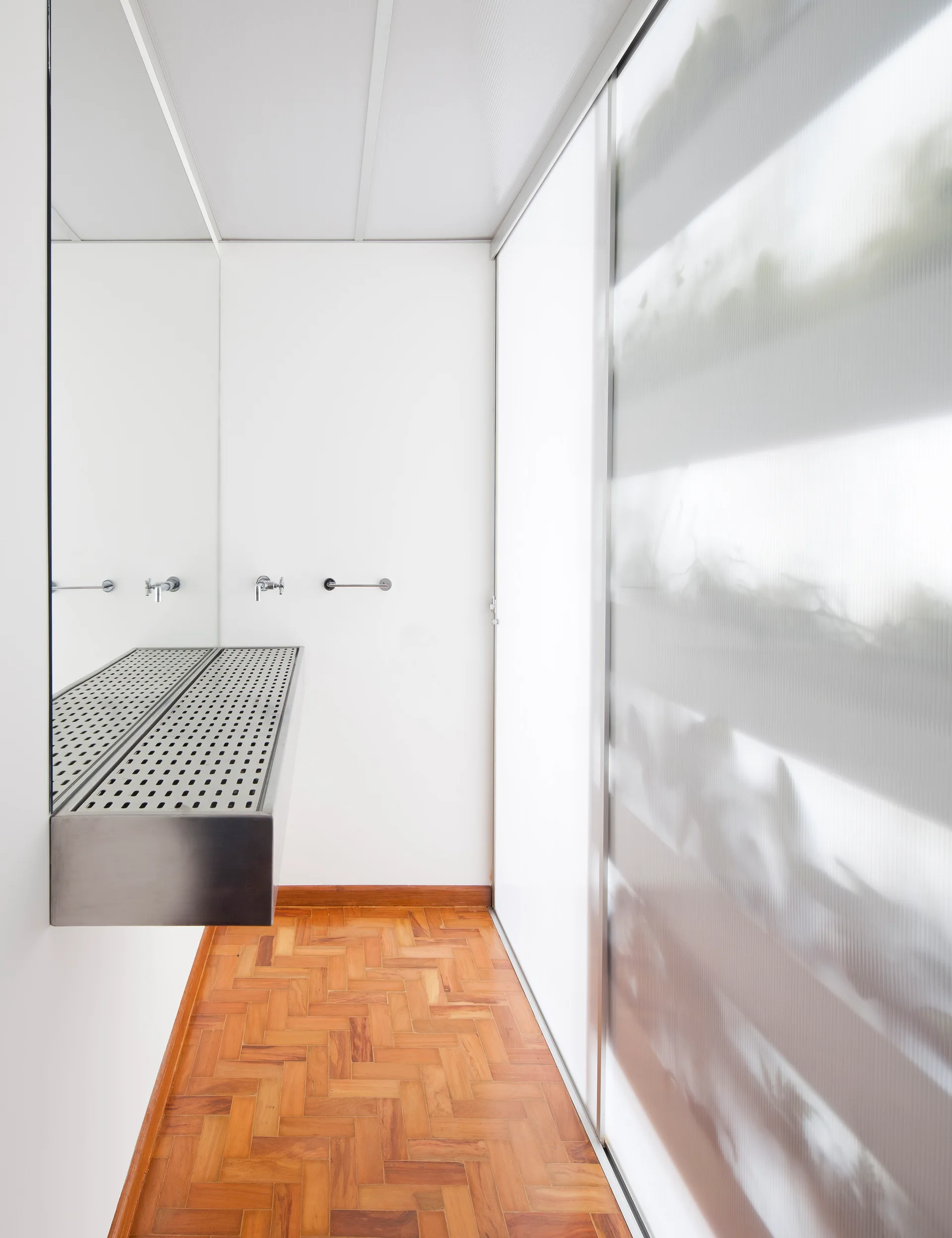 A minimalist white bathroom features a wall-mounted stainless steel vanity, warm parquet wood floors, and a frosted glass sliding door.