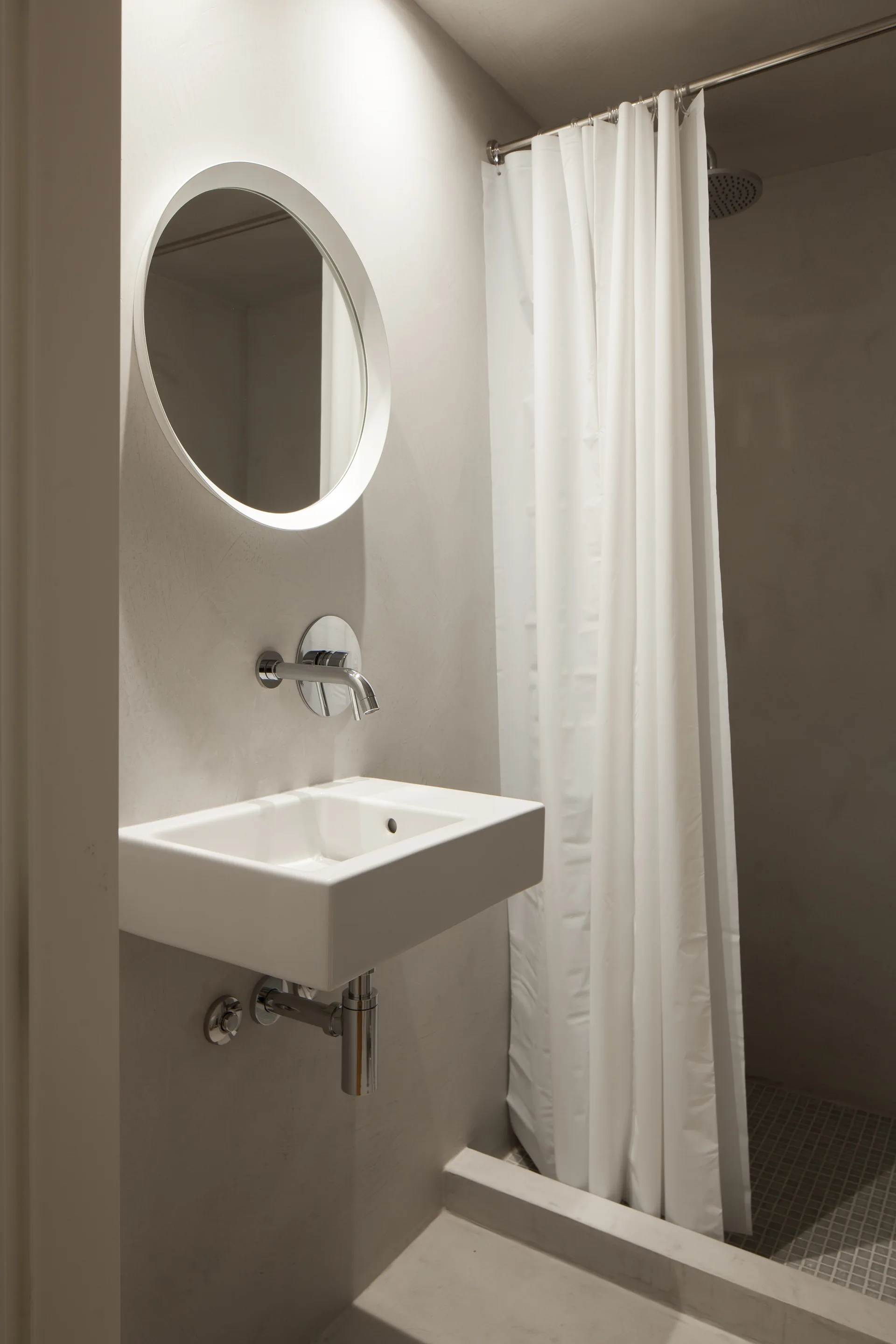 Minimalist bathroom with grey plaster walls, a floating white sink, a circular white-framed mirror, and a white shower curtain.