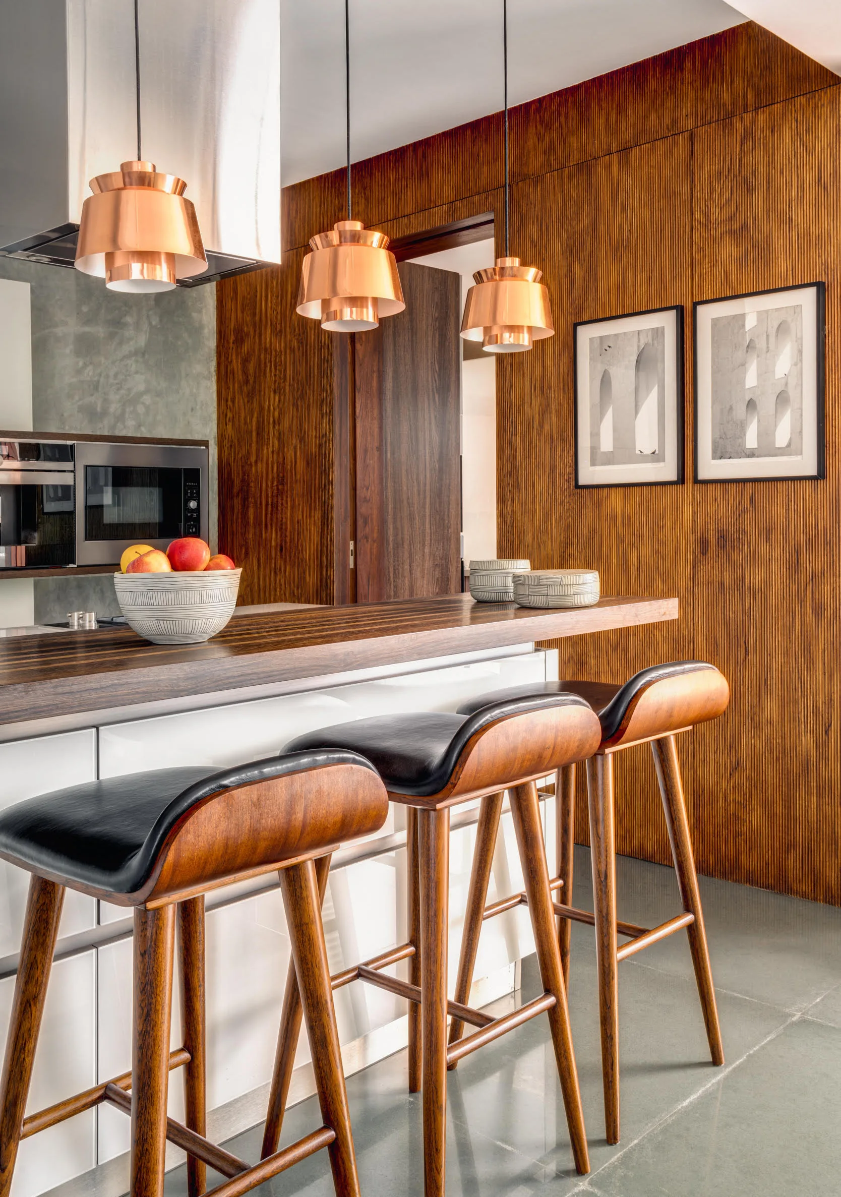 Three wooden bar stools with black leather seats sit at a wood-topped kitchen island beneath three copper pendant lights and a fluted wood wall.