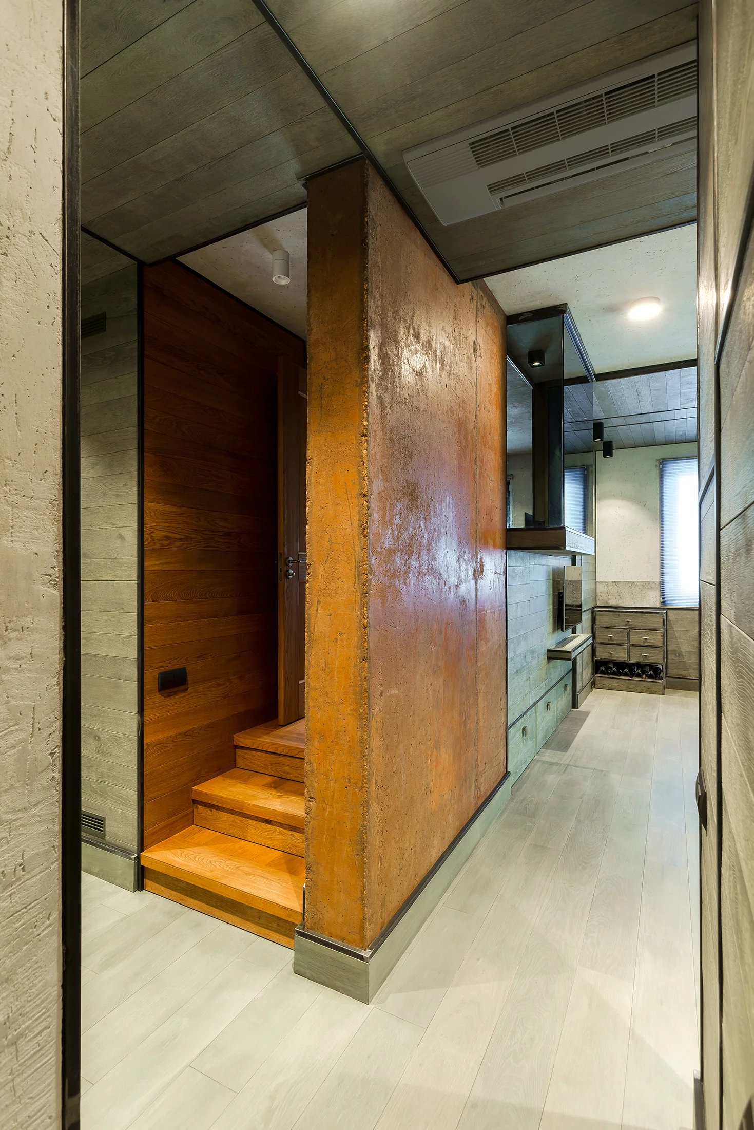 A modern hallway features a central rust-colored partition wall next to a wooden staircase and grey wood-paneled walls.