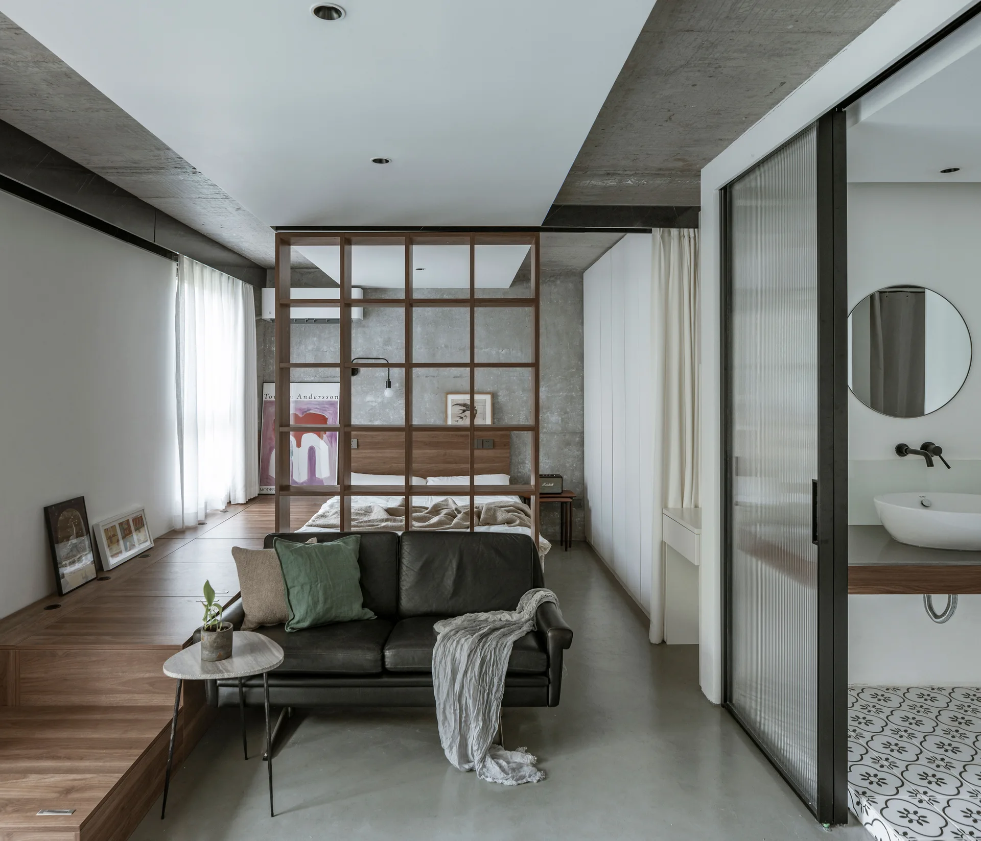 An open-concept studio apartment featuring a black leather sofa, an open wood grid room divider, and a raised wooden platform for the bed.