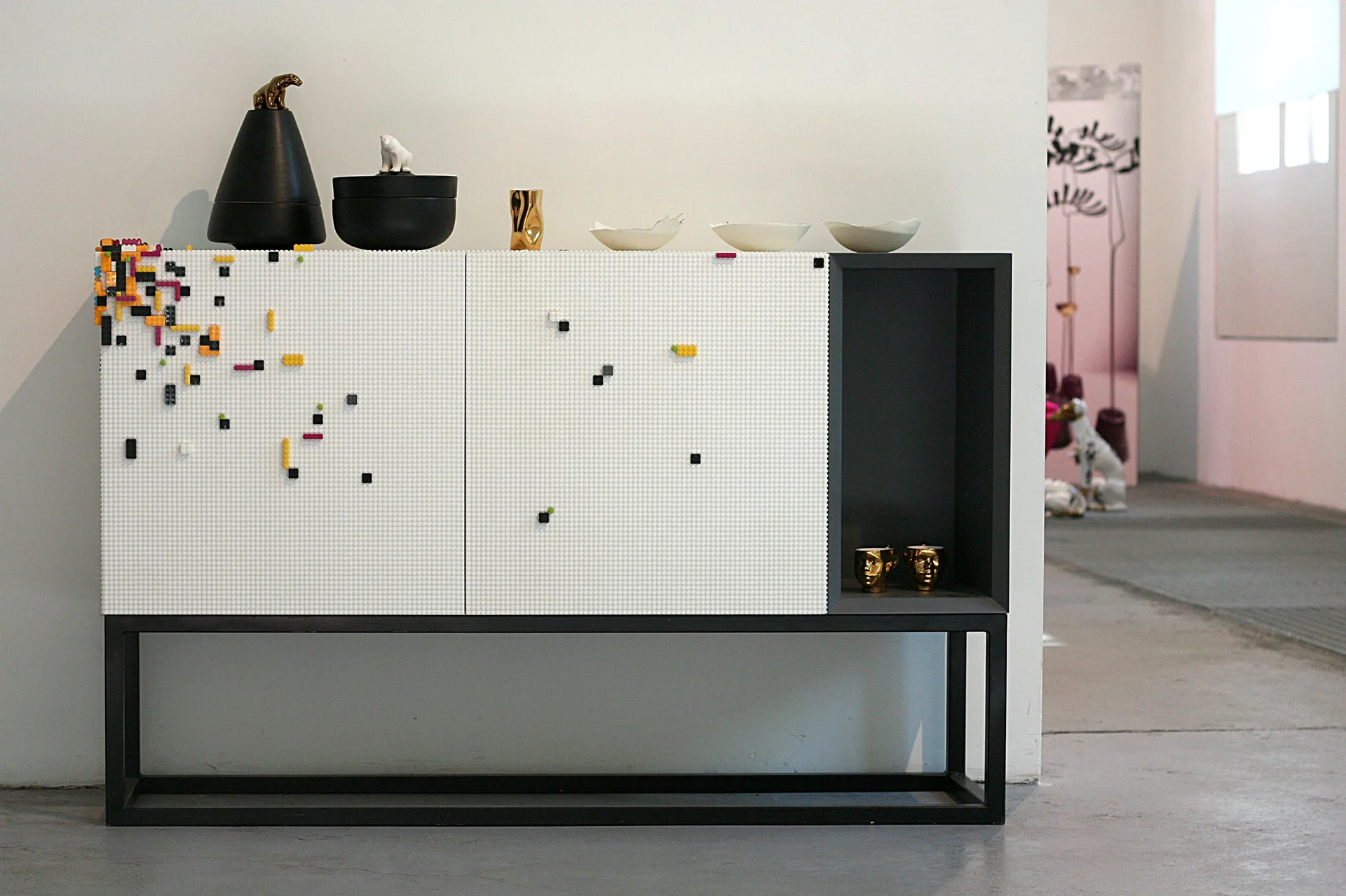 A white storage cabinet with a studded modular brick texture and colorful accents sits on a black metal frame on a concrete floor.