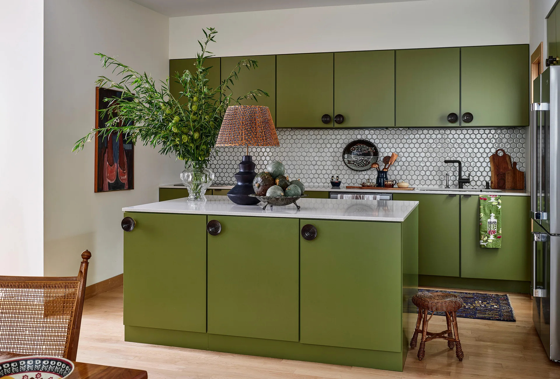 An olive green kitchen featuring matte cabinets with large circular knobs, a white hexagonal tile backsplash, and an island with a stone countertop.