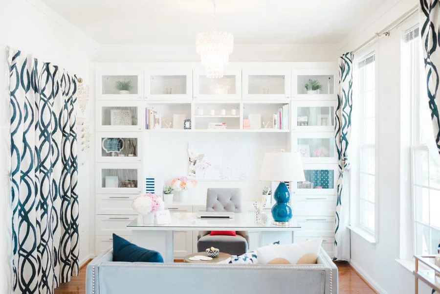A bright home office features a white built-in storage unit, a glass-top desk with a tufted grey chair, and navy patterned curtains.