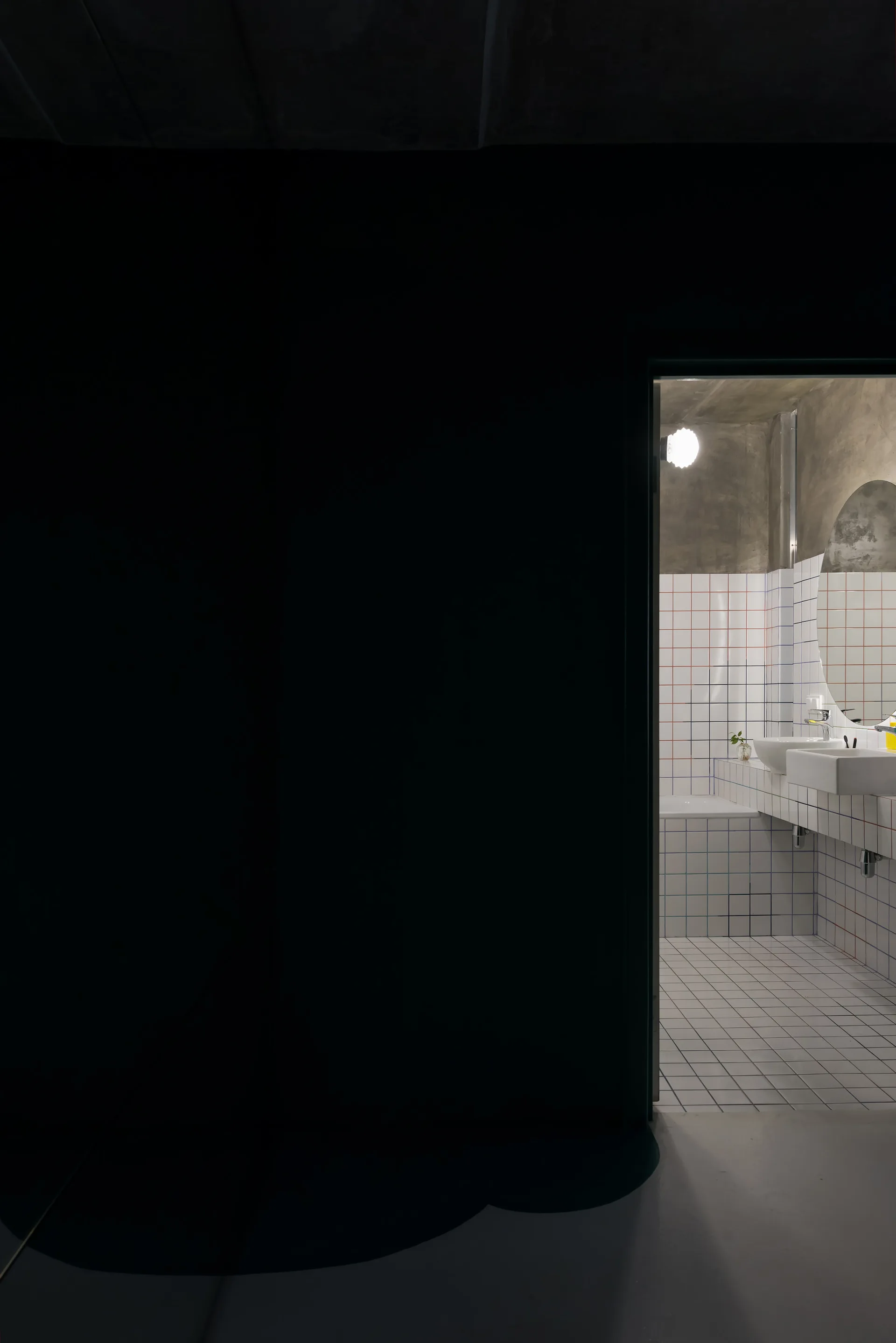 A view through a dark doorway into a bathroom with white grid tiles featuring red and blue lines, a round mirror, and a concrete ceiling.