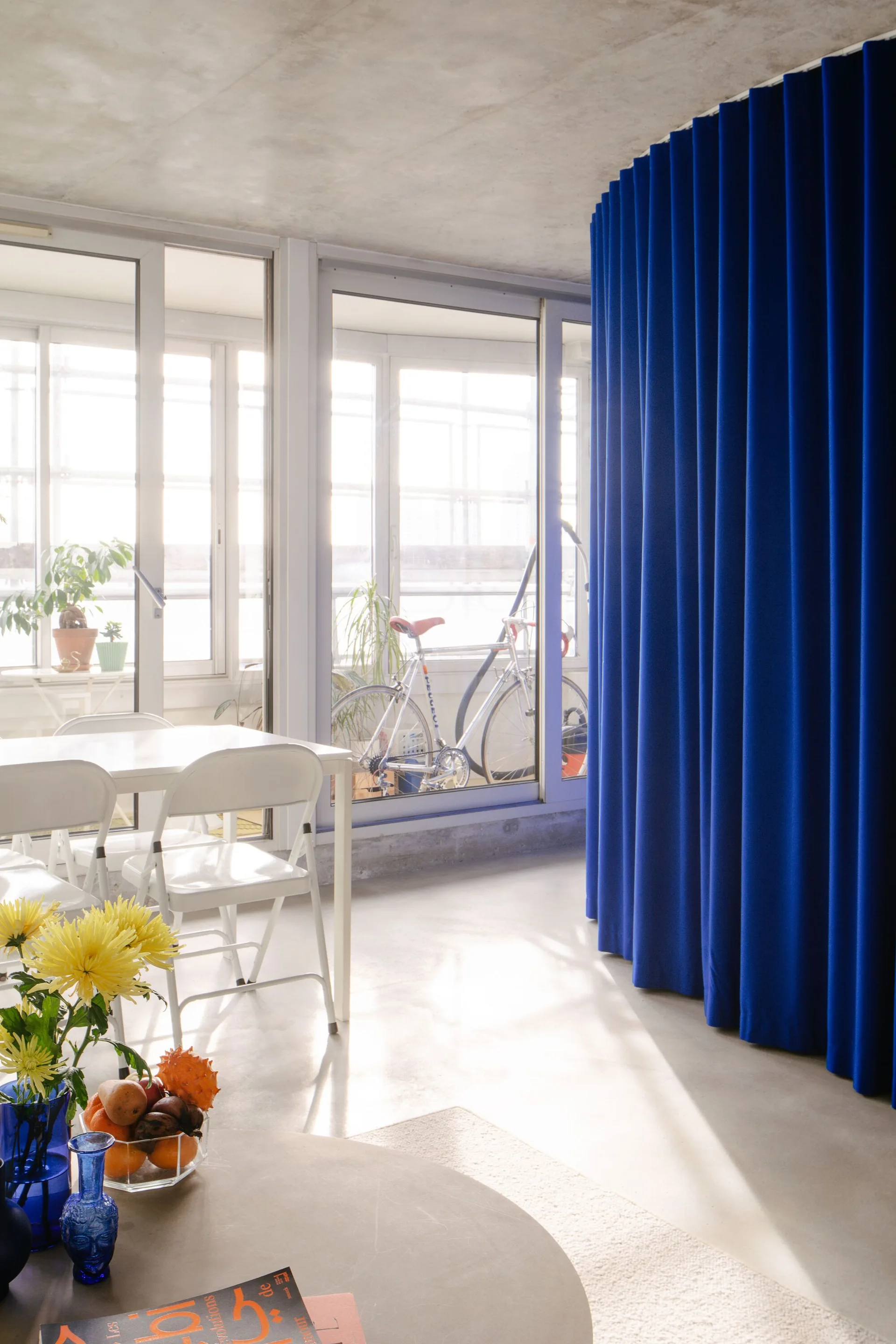 A sunlit dining room with an exposed concrete ceiling, a white table with chairs, and a large curved blue drapery divider.