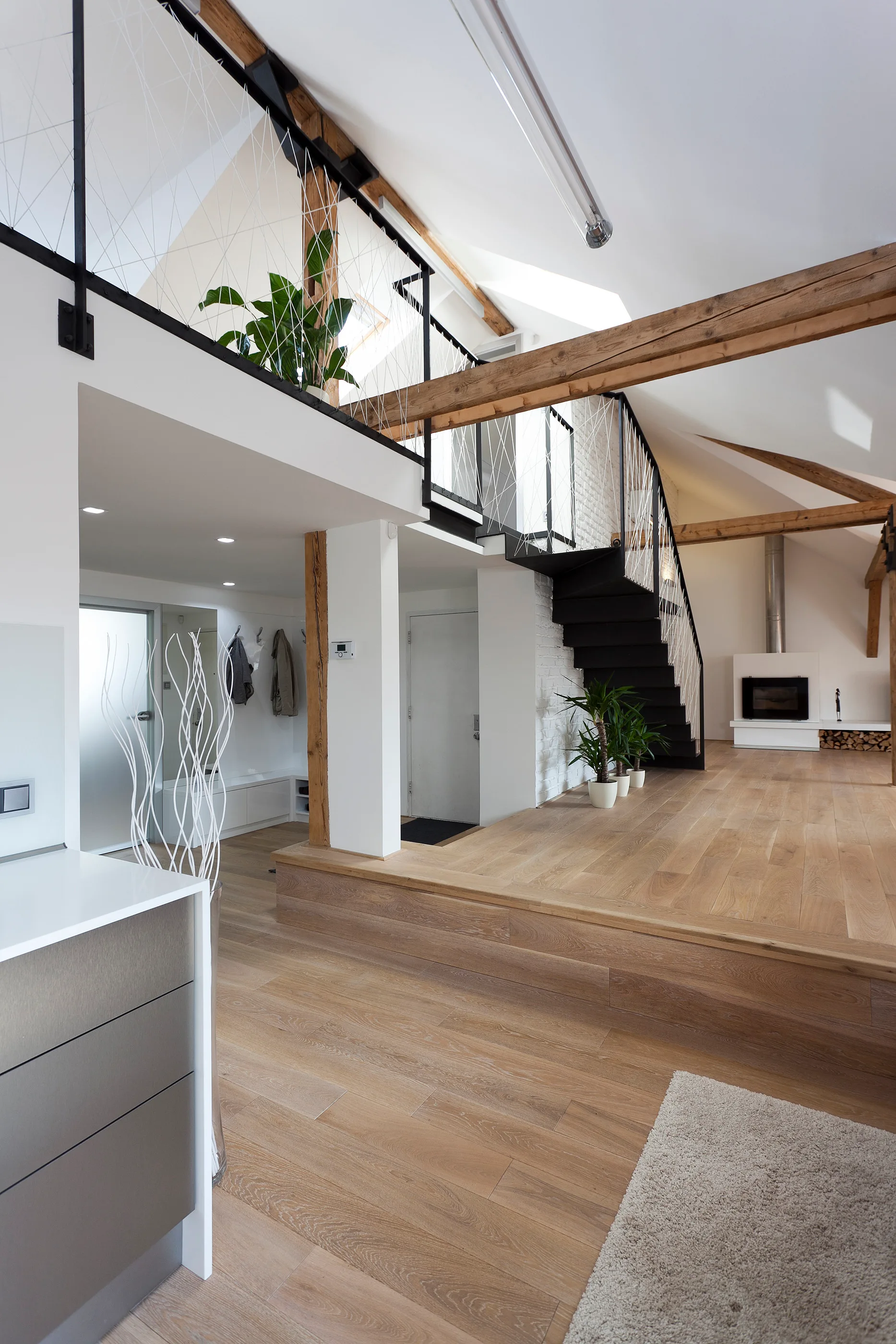 A spacious modern loft features a split-level wooden platform floor, exposed timber beams, a black metal staircase, and a mezzanine with mesh railing.