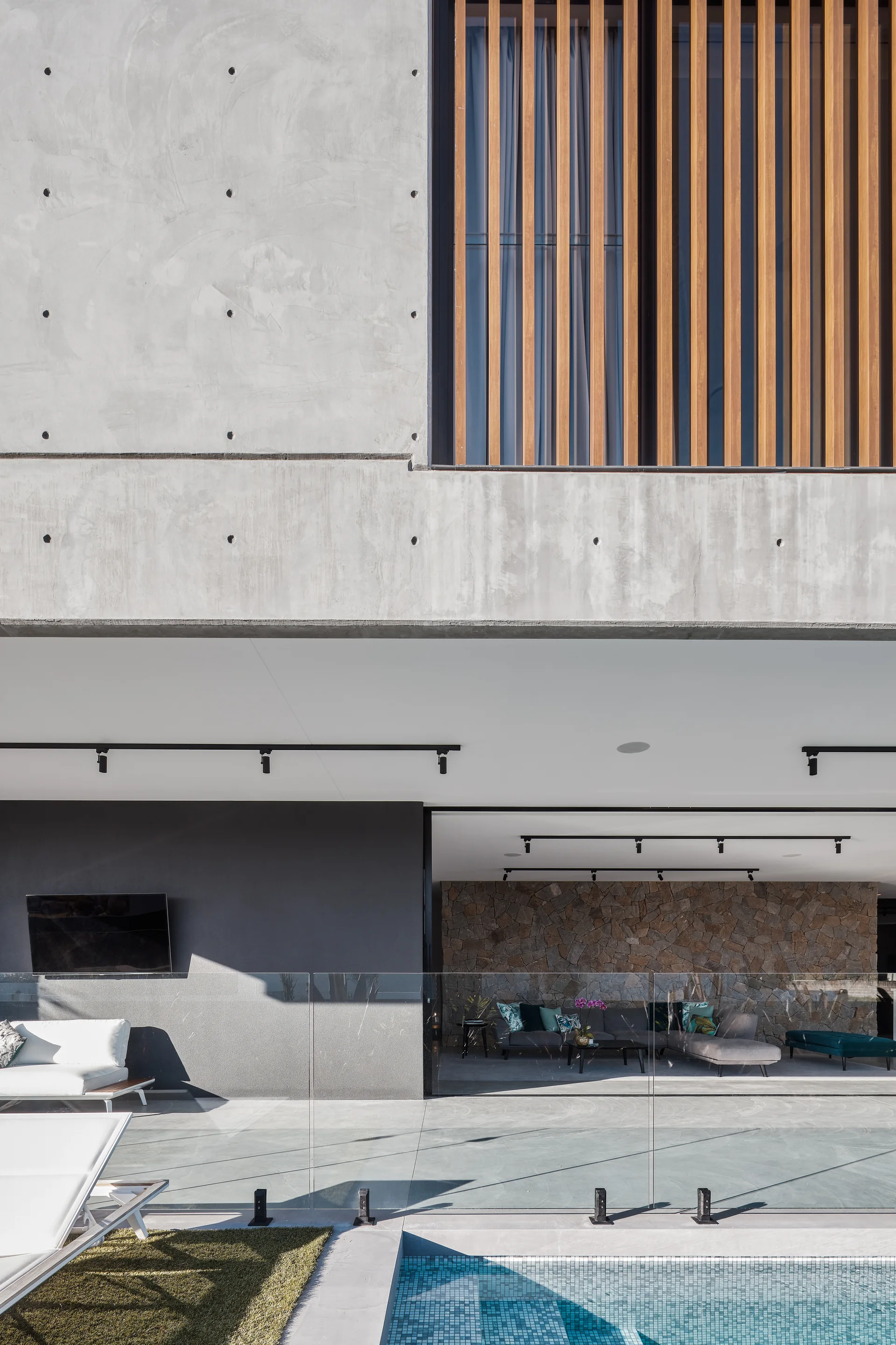 A modern concrete home exterior features a swimming pool, glass railing, and an open-air patio transitioning to an indoor living area with a stone wall.