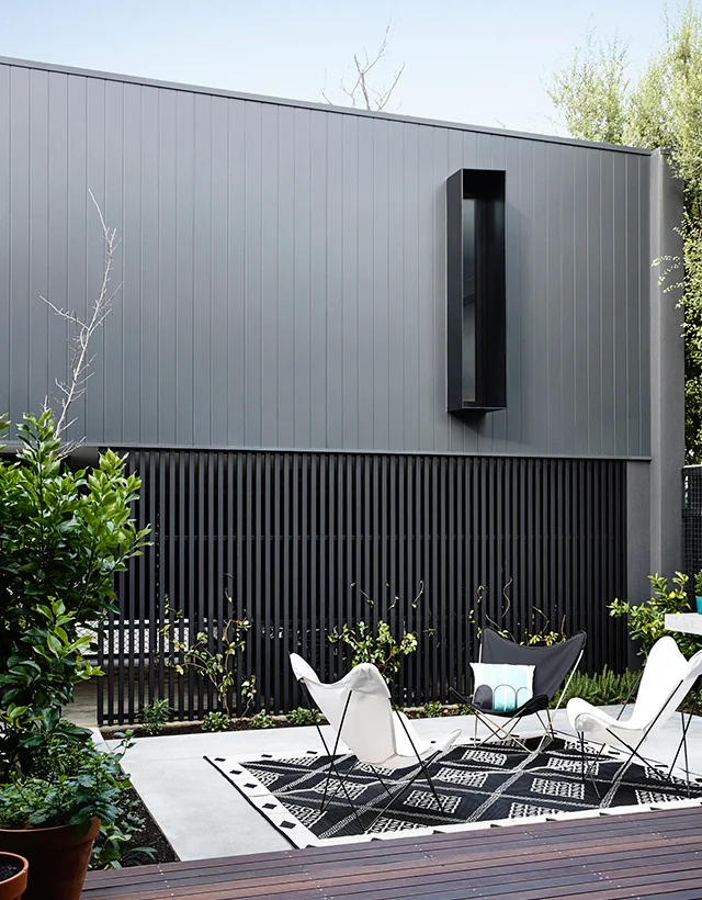Three modern butterfly chairs sit on a black and white geometric rug atop a concrete patio next to a gray vertical-sided house.