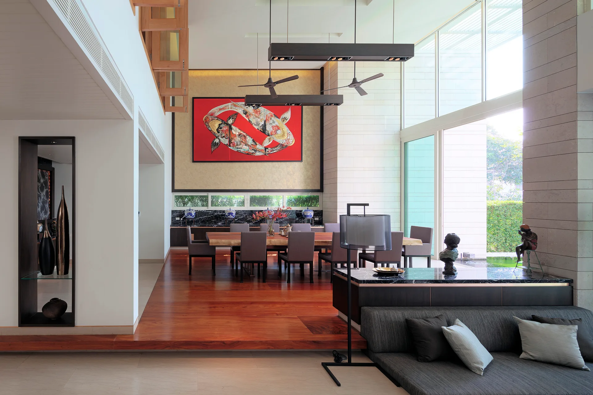 An open-concept living space features an elevated dining table with a red koi painting, a low-profile sofa, and large windows.