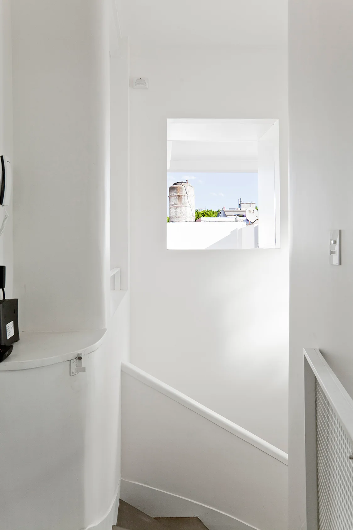 A bright white minimalist stairwell featuring a curved plaster wall and a square window that looks out onto a rooftop.