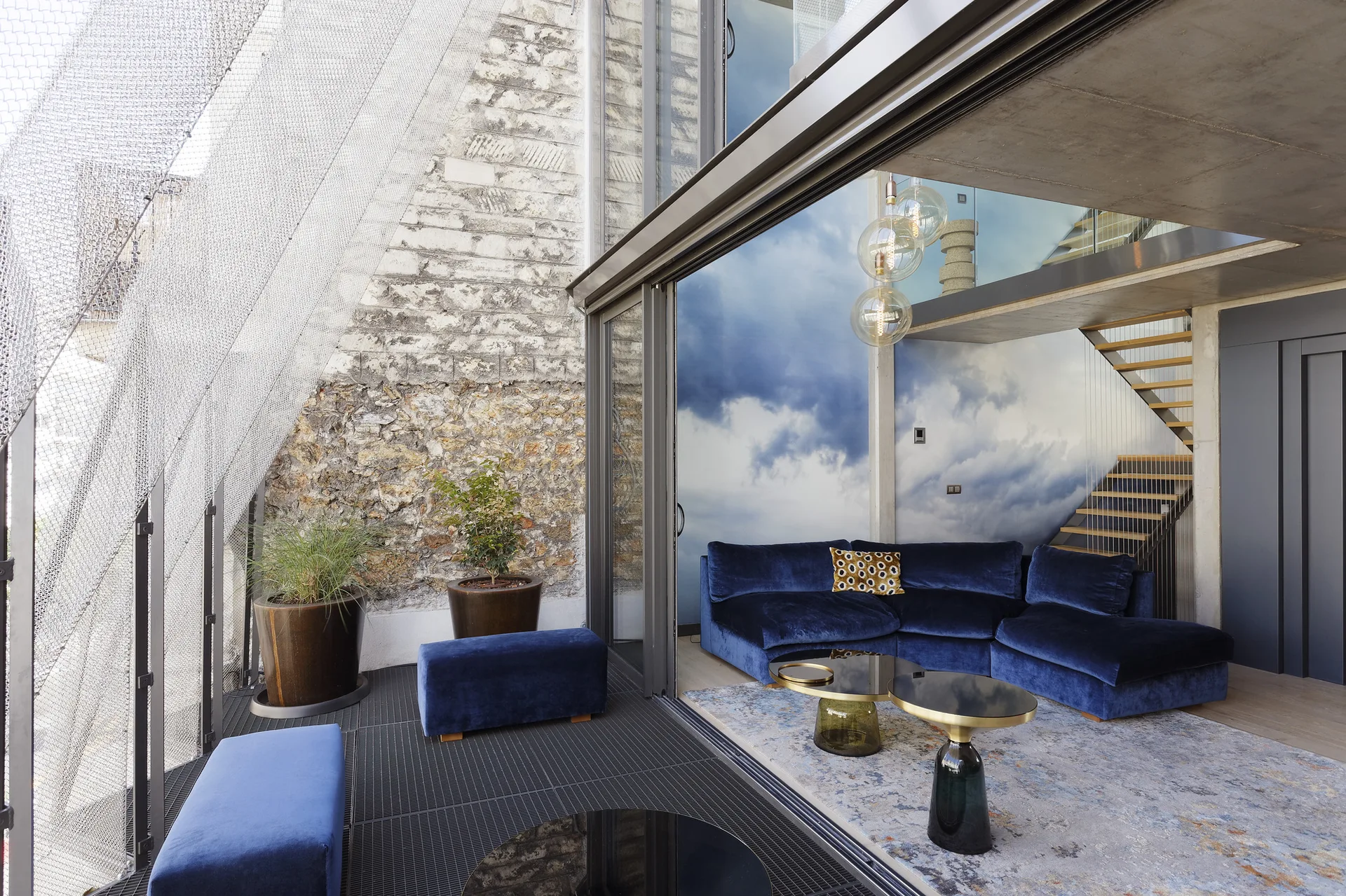A modern living room features a blue velvet sectional sofa, a cloud-themed wall mural, and sliding glass doors that open to an outdoor terrace.