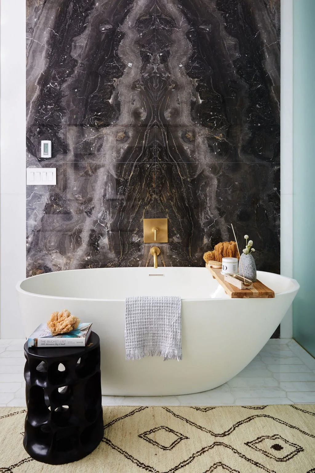 A white freestanding bathtub sits against a dark veined marble feature wall accented by a gold-tone wall faucet and a black sculptural side table.