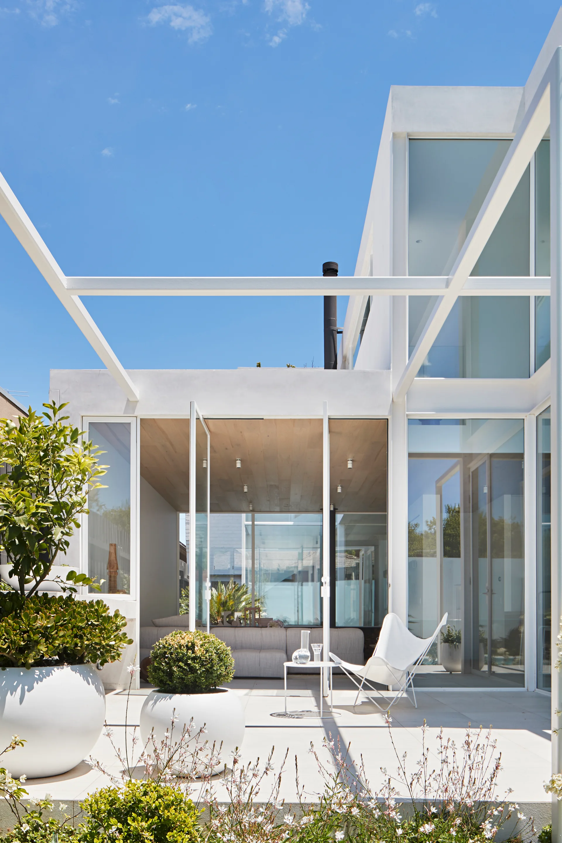 A minimalist white patio features large spherical planters, a butterfly chair, and an overhead pergola frame against a modern house facade.