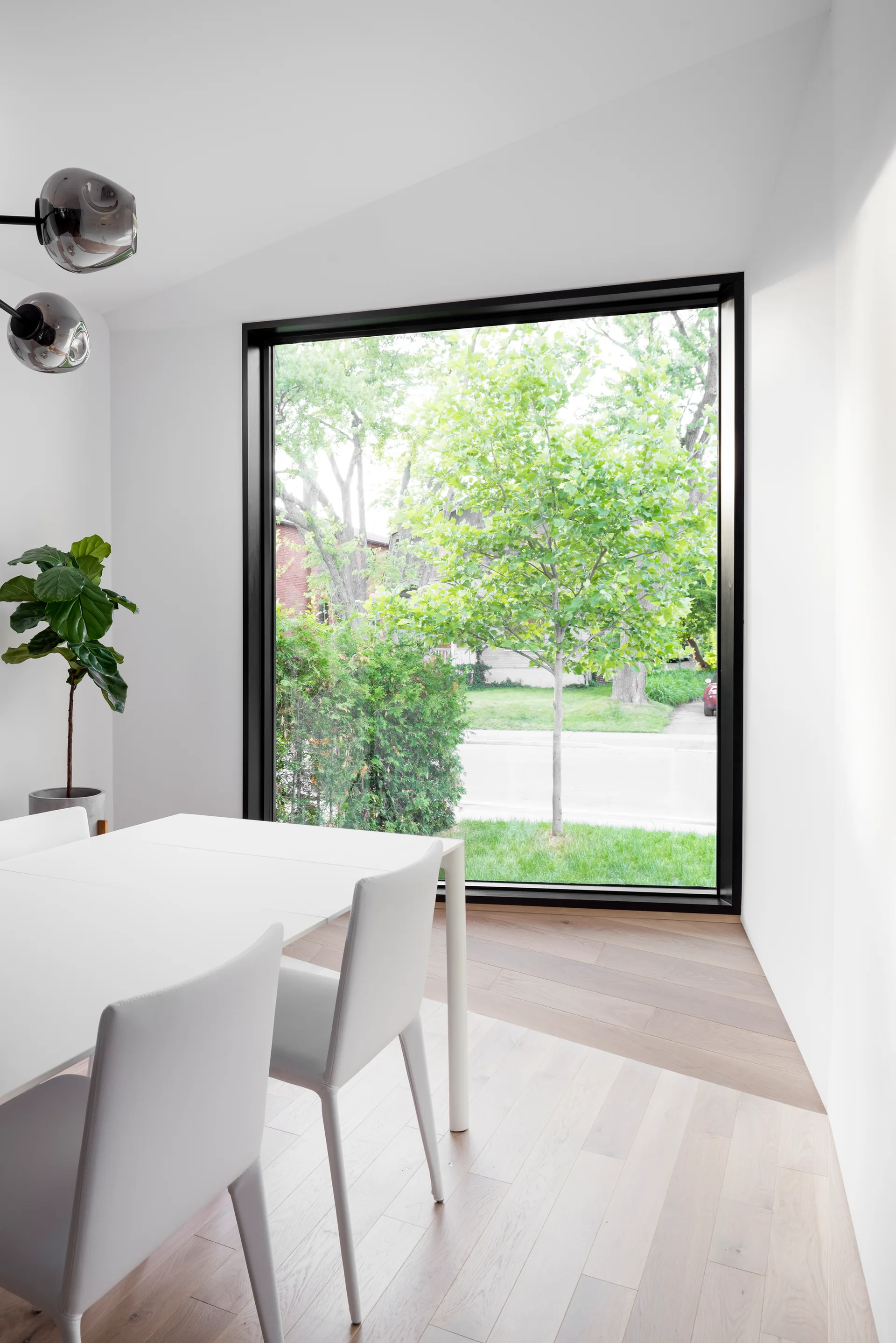 A minimalist white dining table and matching chairs sit next to a large black-framed picture window looking out onto a green tree.