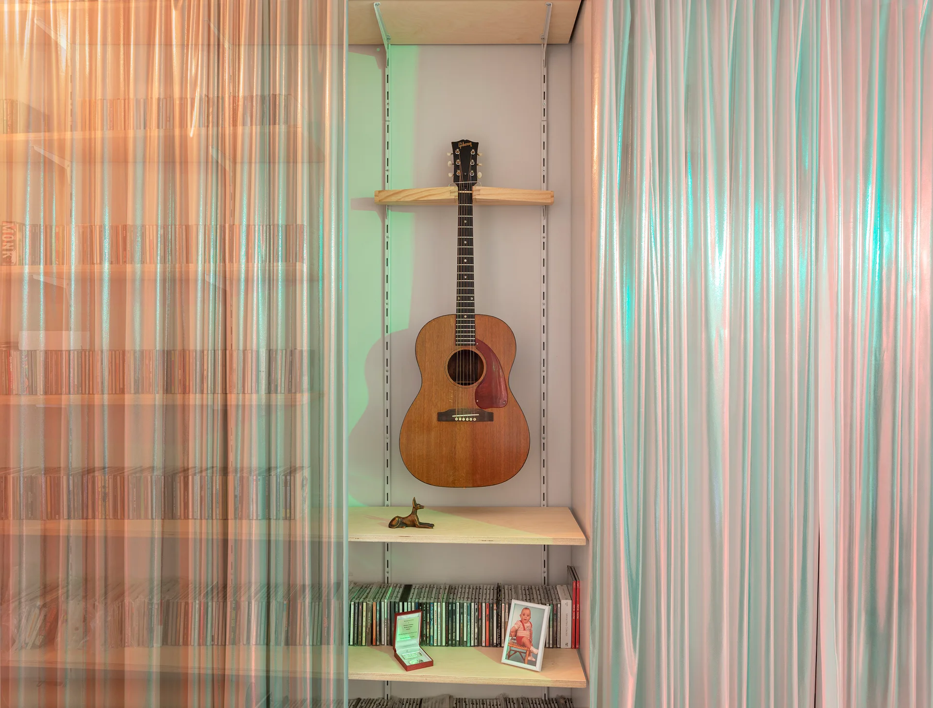 An acoustic guitar hangs on light wood shelves flanked by shimmering iridescent curtains reflecting orange and green hues.