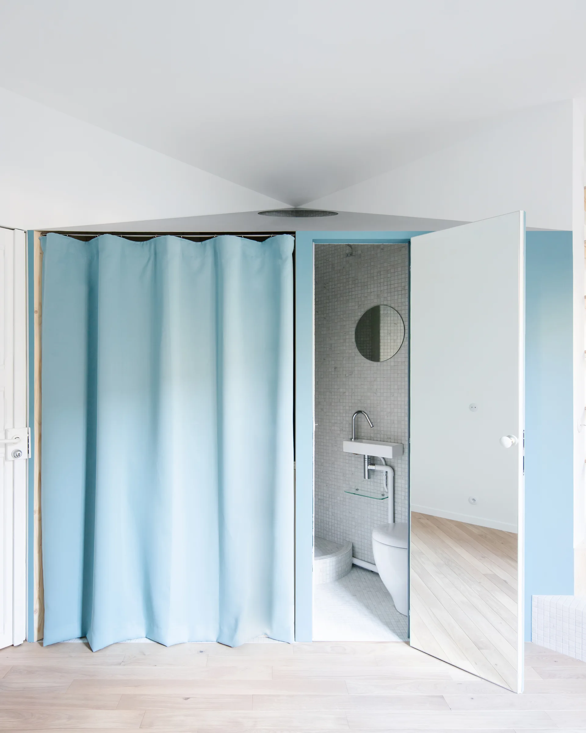 An open white door with a full-length mirror reveals a small bathroom with mosaic tiles, next to a large light blue curtain hanging from a matching frame.