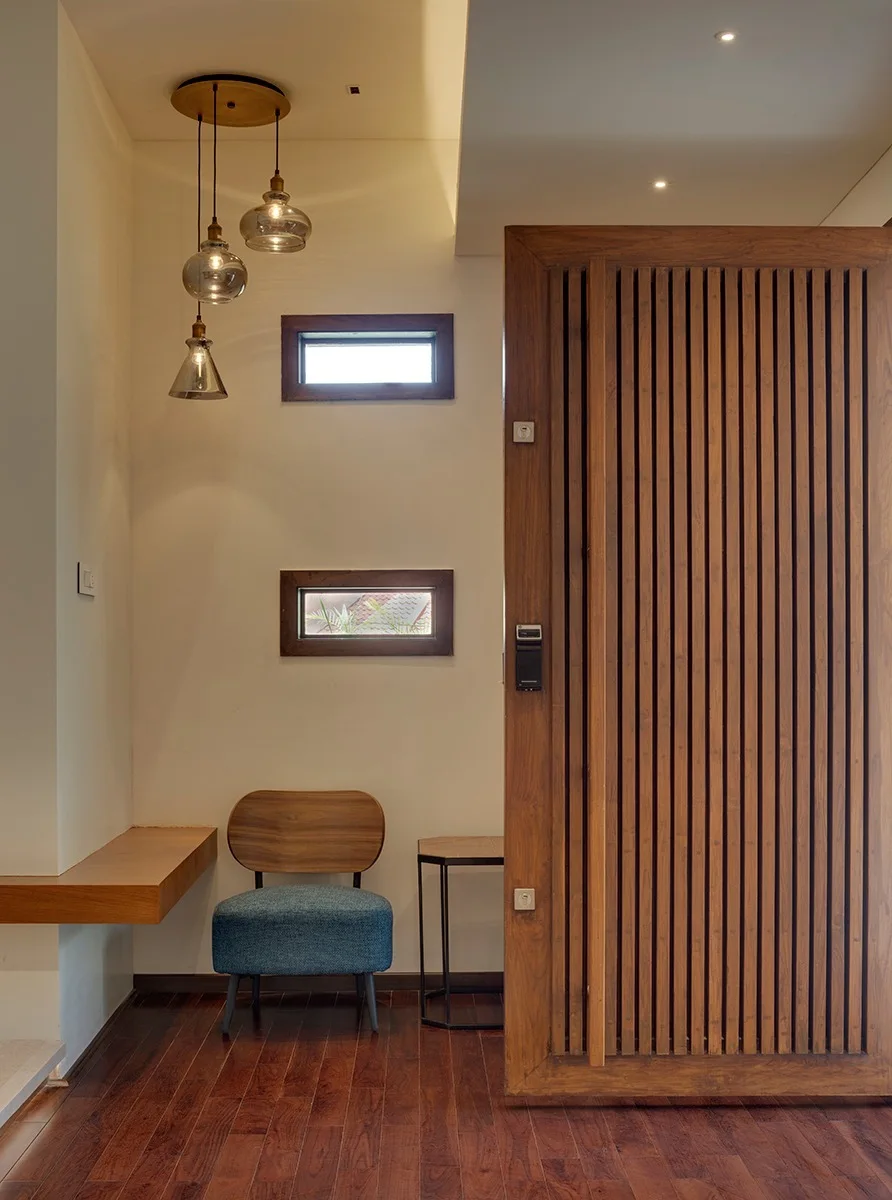 An entryway features a large slatted wooden door, a blue accent chair, and cluster pendant lights hanging beneath two small horizontal windows.