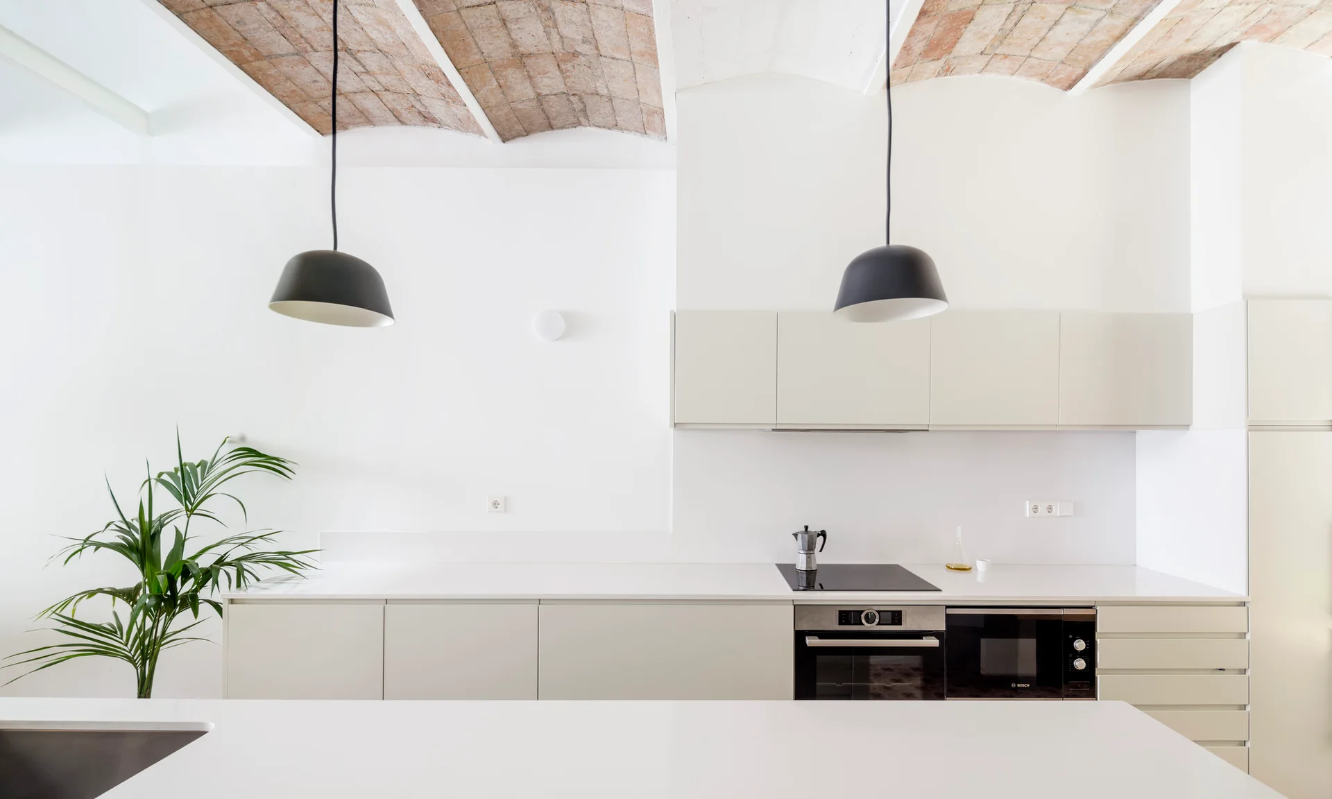 A minimalist kitchen features flat-front beige cabinets, white countertops, black dome pendant lights, and a vaulted brick ceiling.