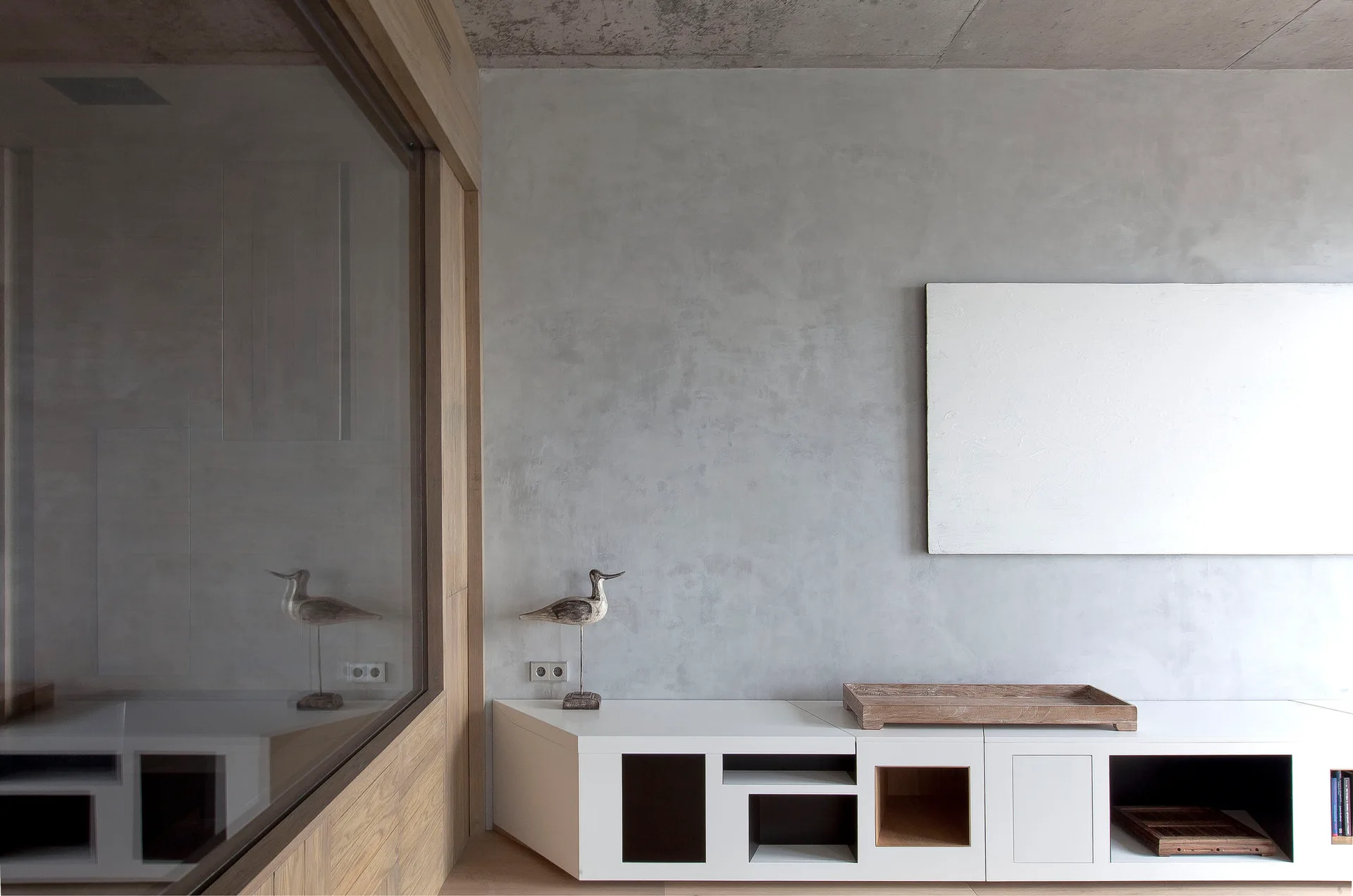 A minimalist living space features a low white media console against a textured grey plaster wall next to a wood-framed glass partition.