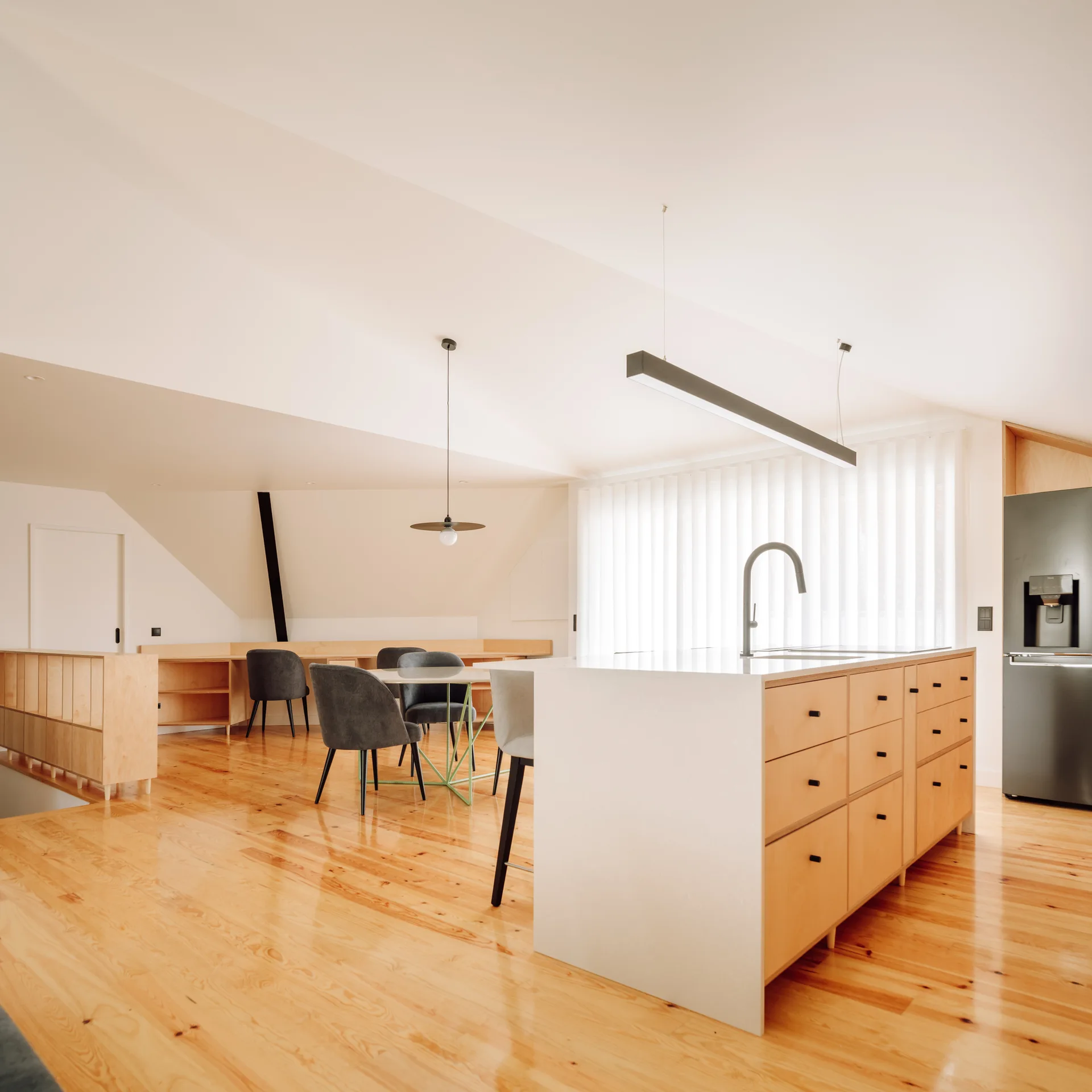 A modern open-plan kitchen featuring a blonde wood island with white countertops, polished wood floors, and grey dining chairs under an angled ceiling.