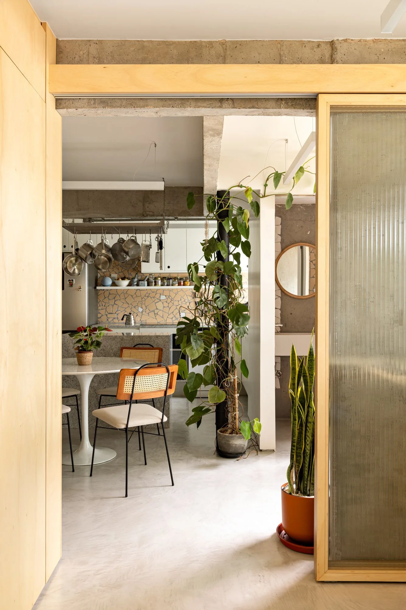 A dining area viewed through a doorway features a white tulip table with cane-back chairs, a fluted glass door, and exposed concrete ceilings.