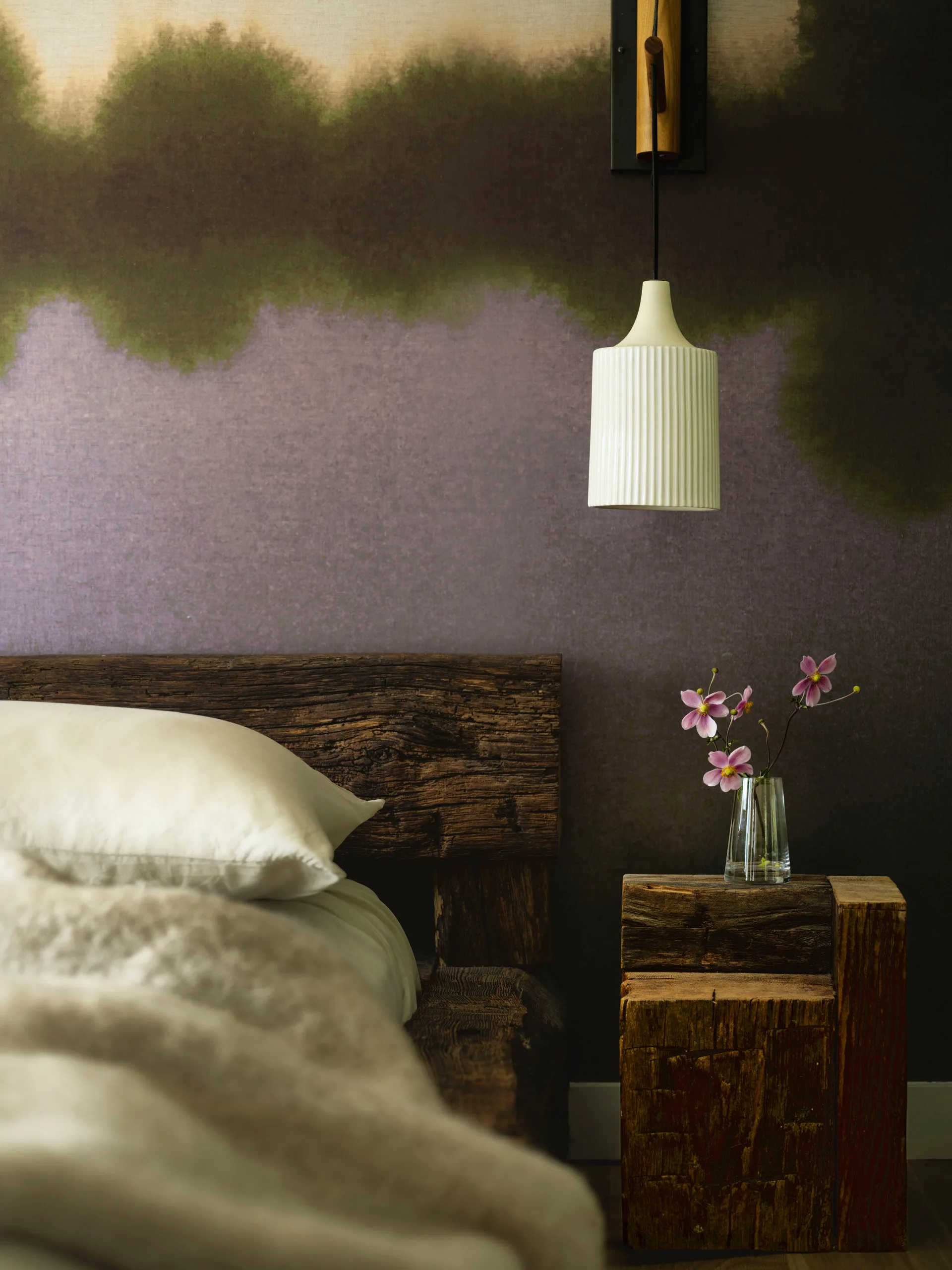 A rustic wood headboard and block nightstand sit against purple and brown landscape mural wallpaper next to a fluted white pendant light.