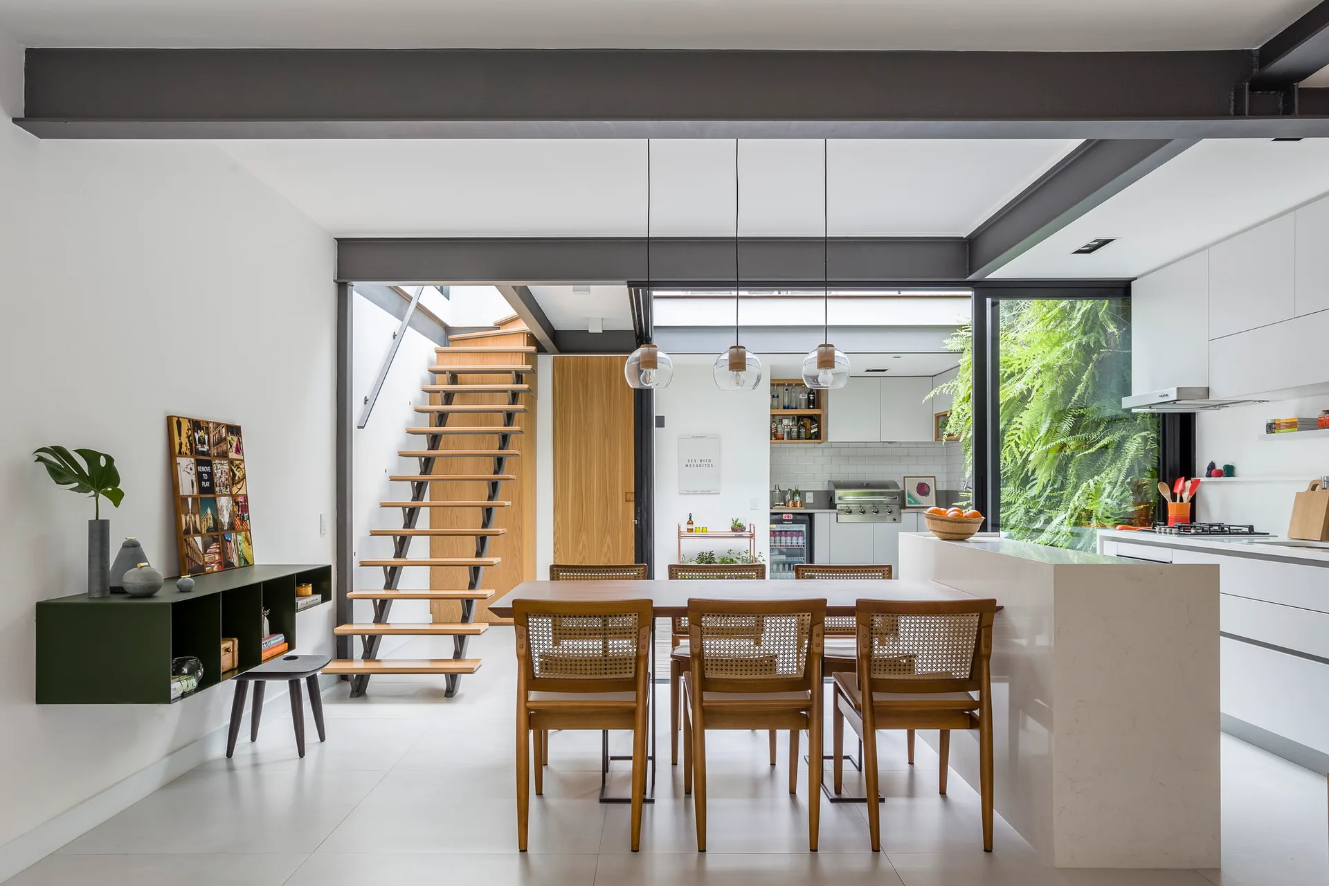 A modern open-concept dining room featuring a wooden table with cane chairs, a floating wood staircase, and black steel structural beams.