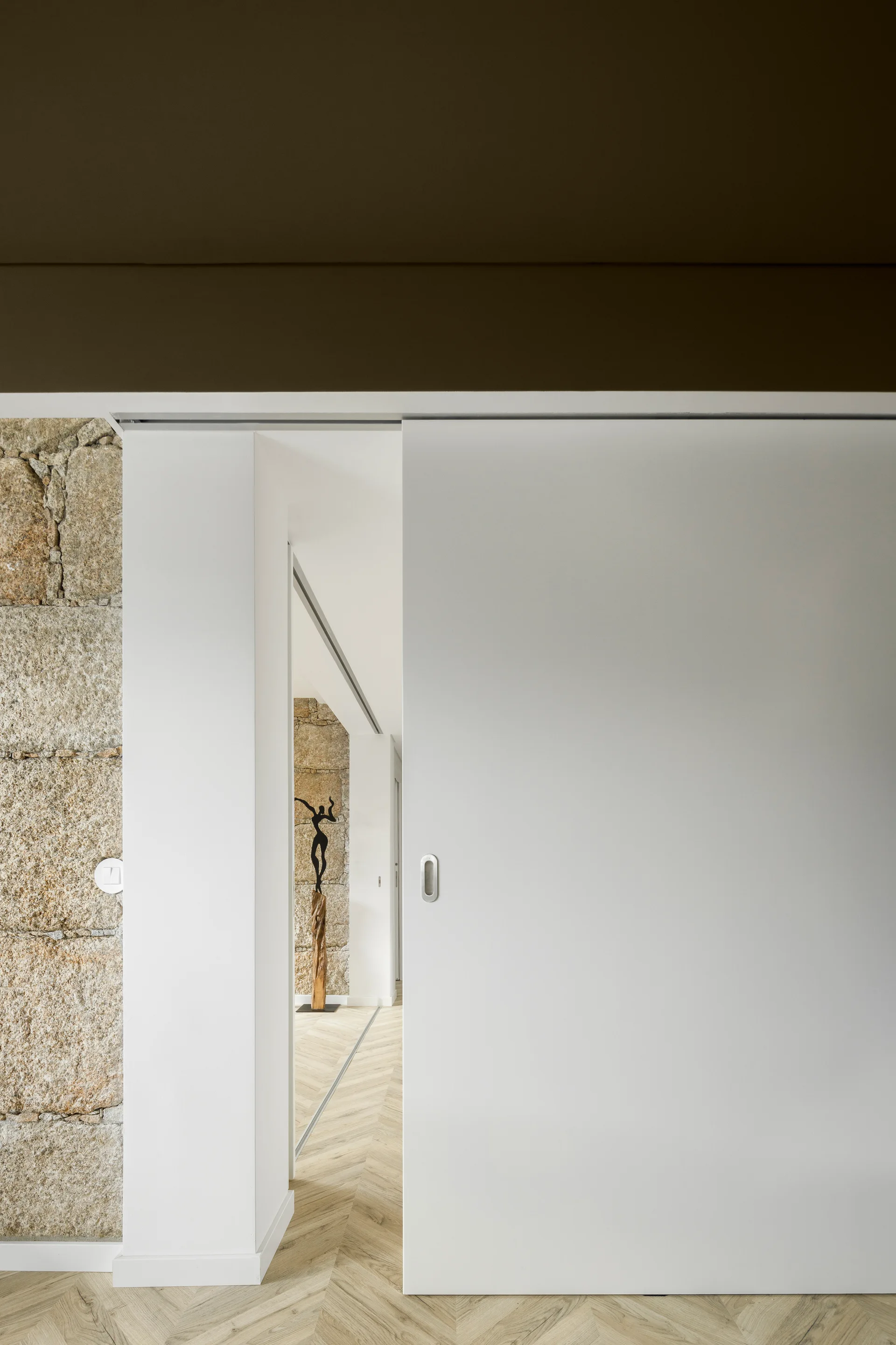 A modern hallway features a white sliding door, an exposed stone wall, and light wood herringbone flooring.
