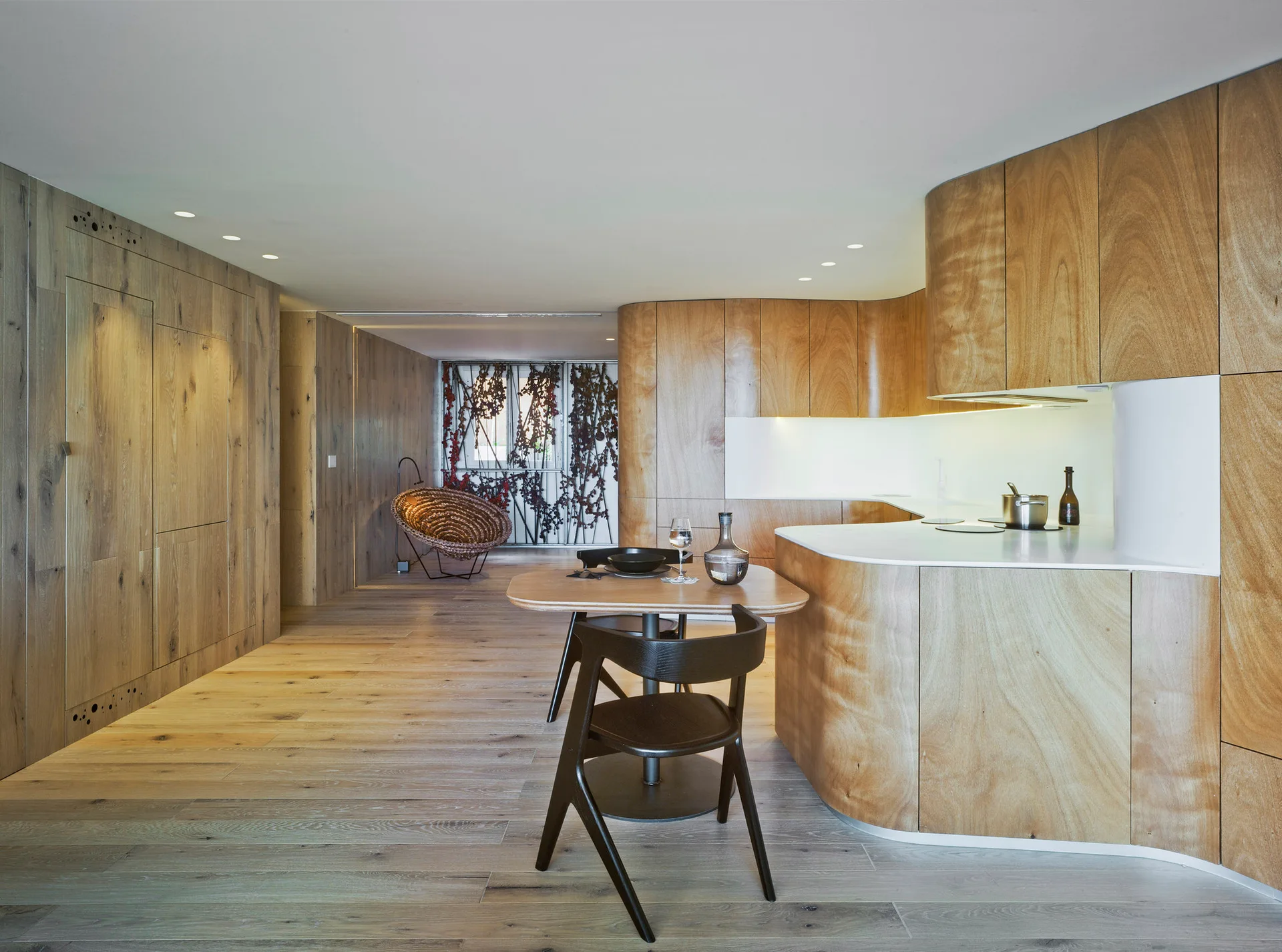 A minimalist open-plan kitchen featuring curved light wood cabinetry, matching wall paneling, a small dining table with dark chairs, and recessed lighting.