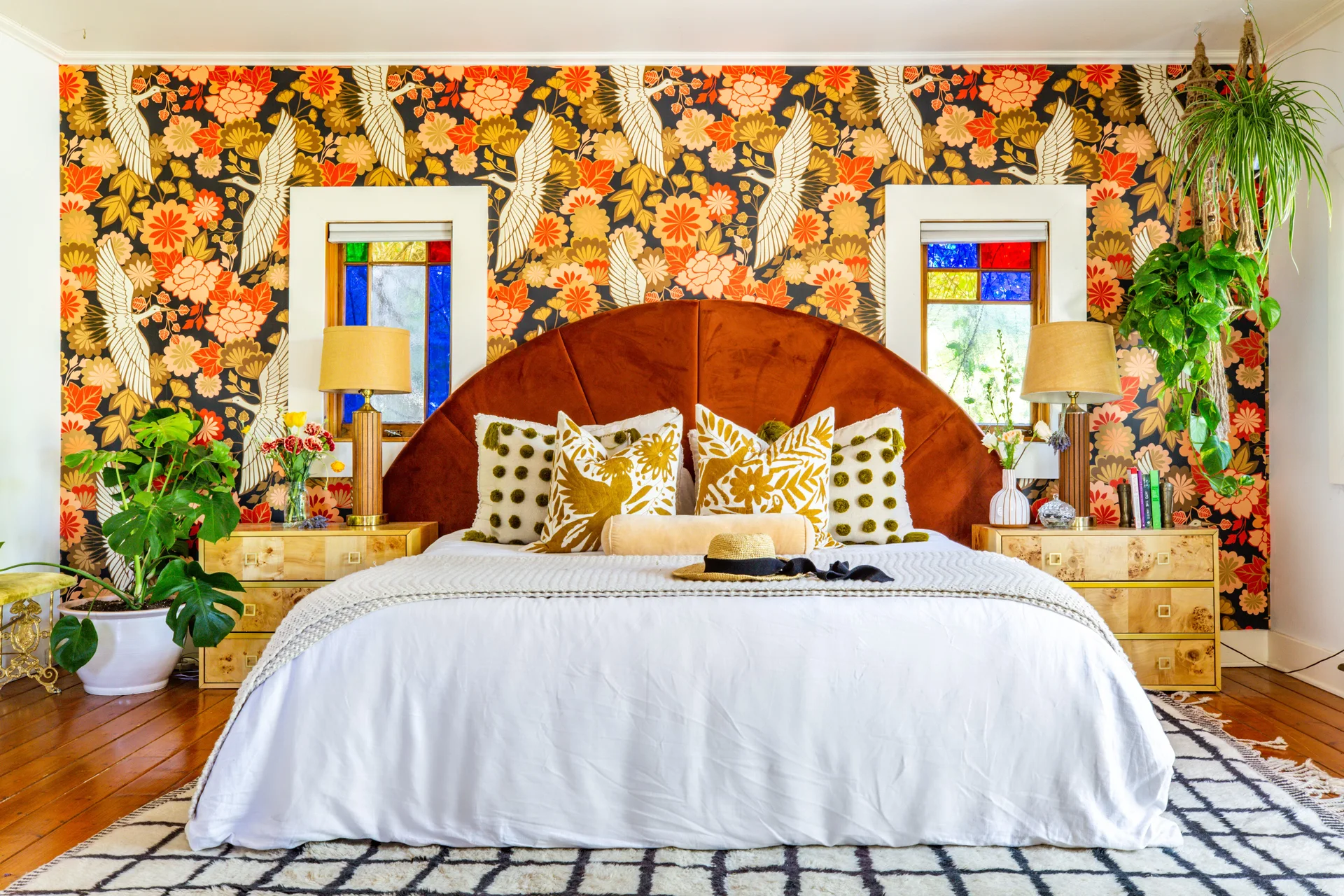 An eclectic bedroom features a bed with a rust-colored headboard, crane wallpaper, and burl wood nightstands flanked by stained glass windows.