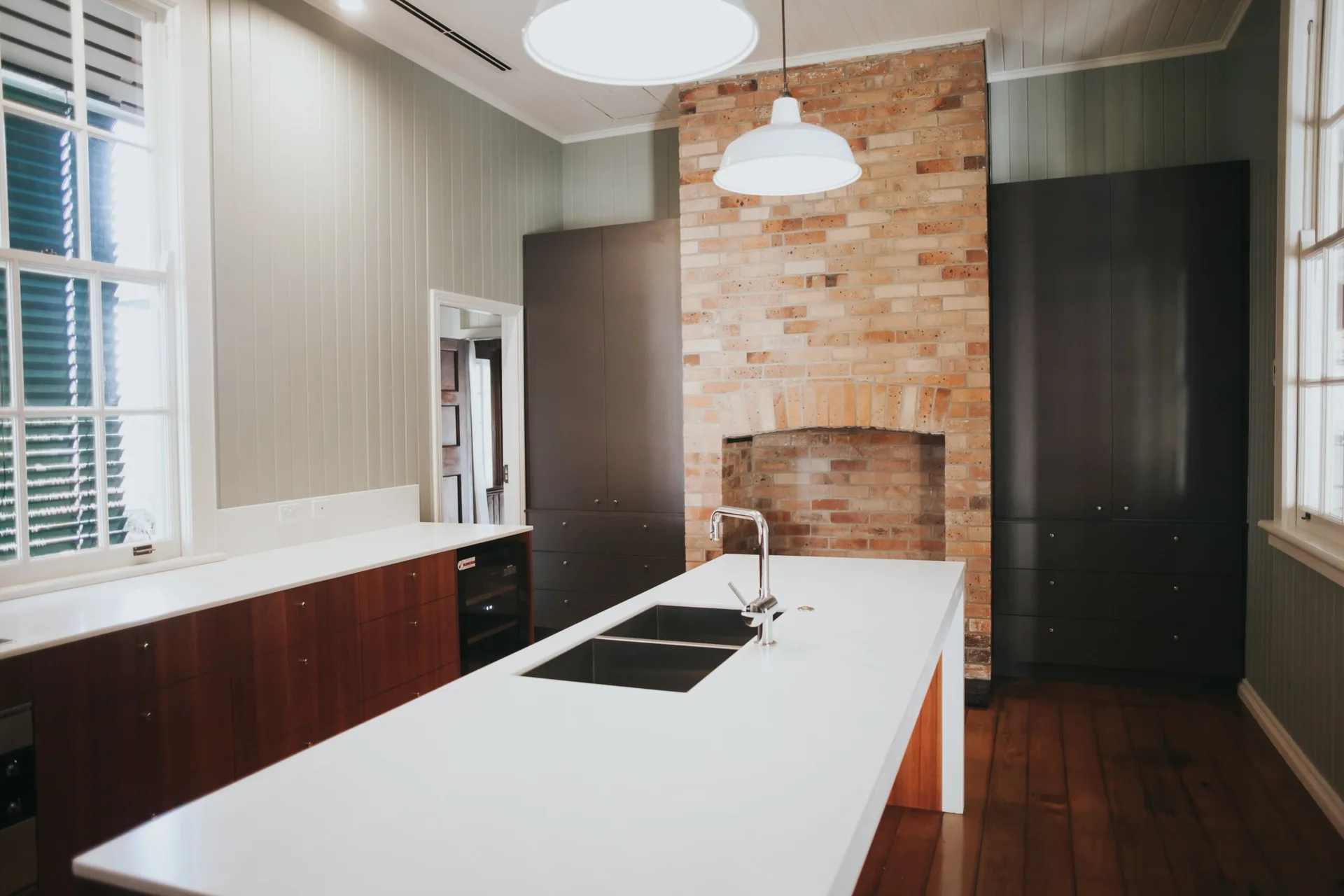 A modern kitchen with a large white island, sage green vertical wall paneling, dark grey cabinets, and an exposed brick fireplace.