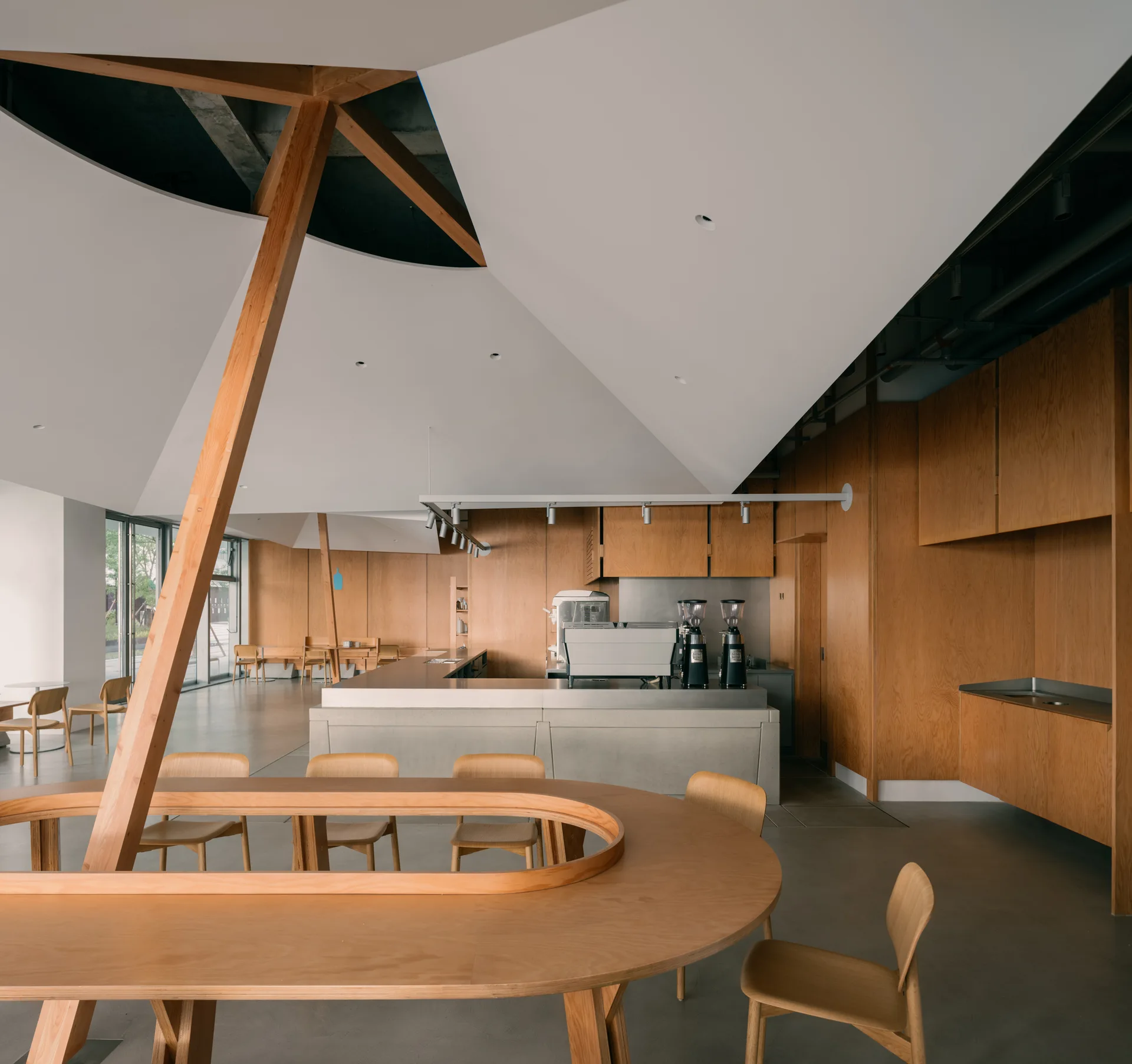 A minimalist cafe interior featuring a large wooden communal table, a concrete counter with espresso machines, warm wood paneling, and an angular white ceiling.