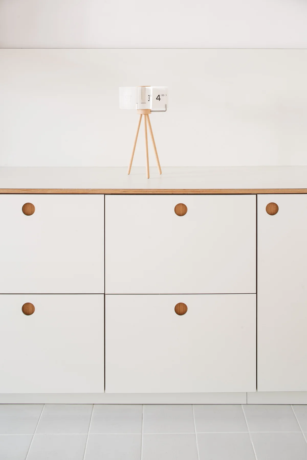 A white tabletop flip clock on a wooden tripod stand sits on a white storage cabinet with circular recessed wooden handles.