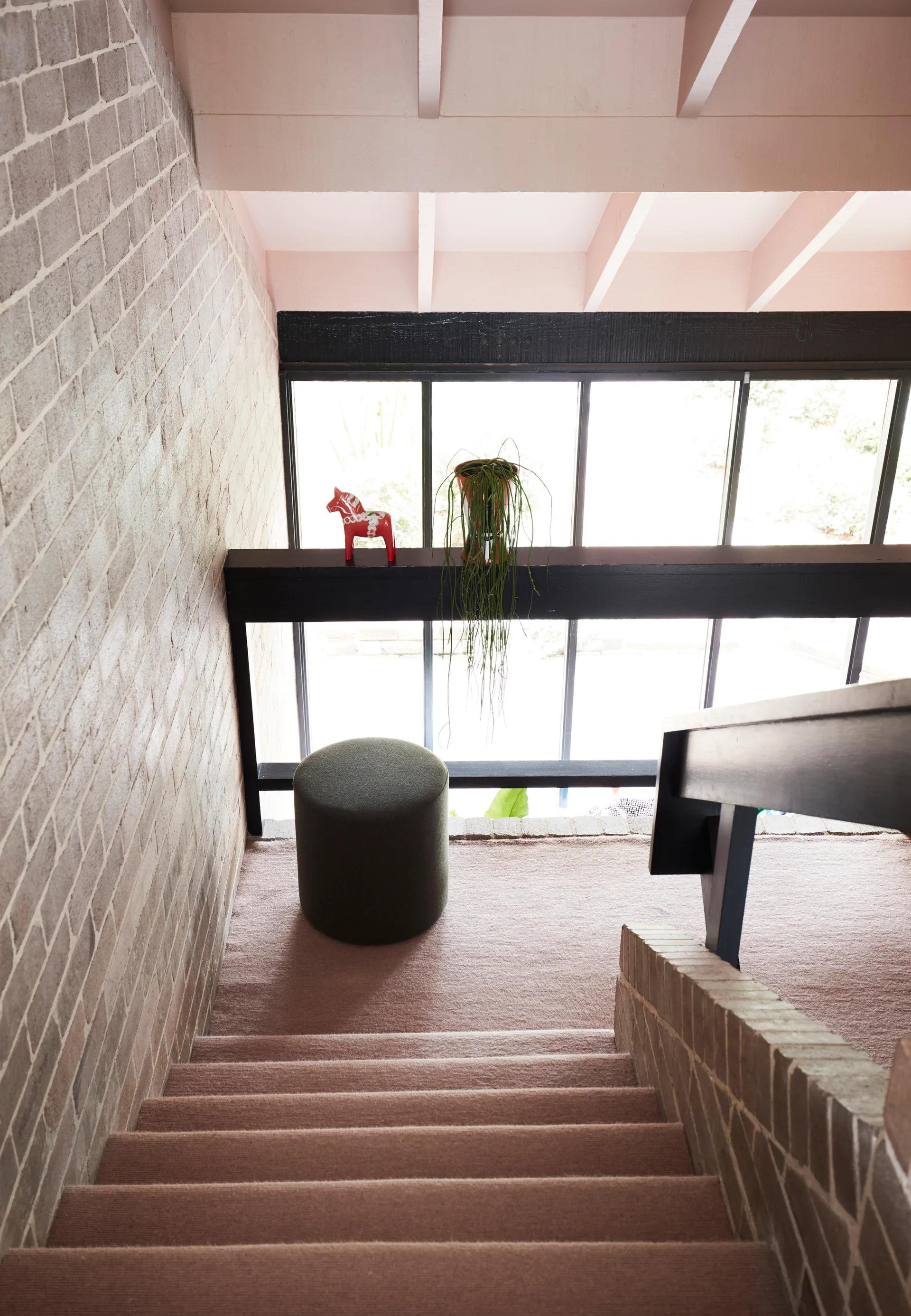 A staircase with dusty pink carpet and a light brick wall leads down to a landing with a green ottoman, black railings, and a large window.