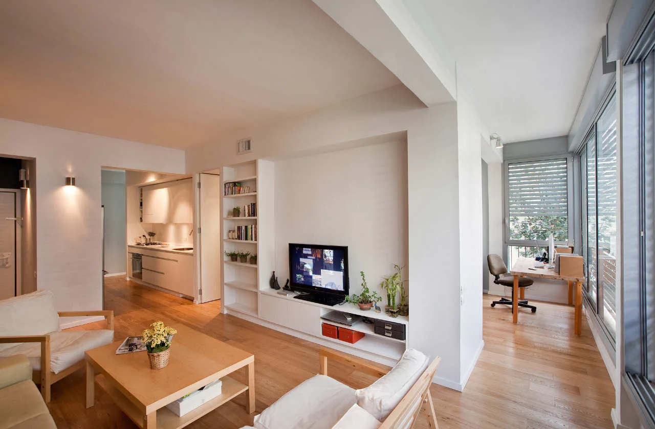 A bright open-plan living room with light wood floors, a white built-in media console, a wooden coffee table, and an adjacent window study nook.